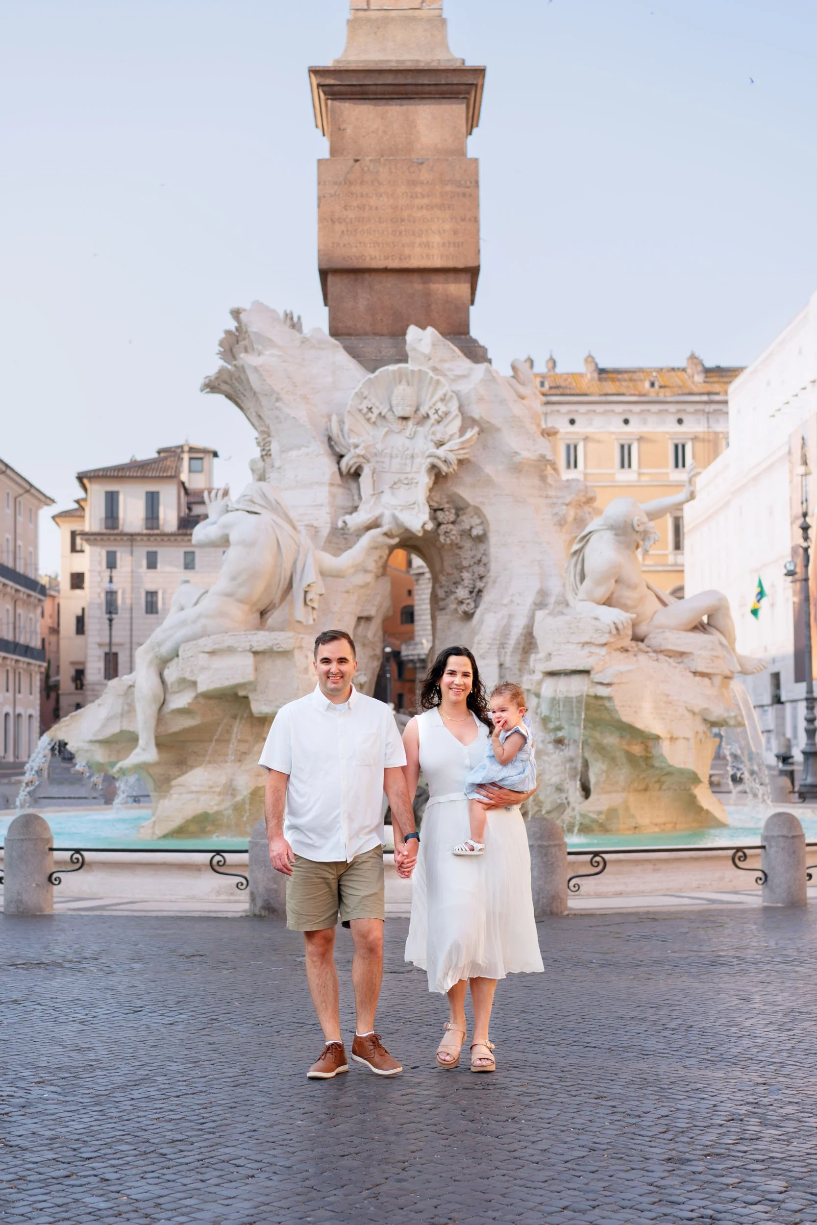 A family of three standing in front of the Fountain of the Four Rivers in Rome, Italy, during daytime. The fountain features four statues representing four major rivers.