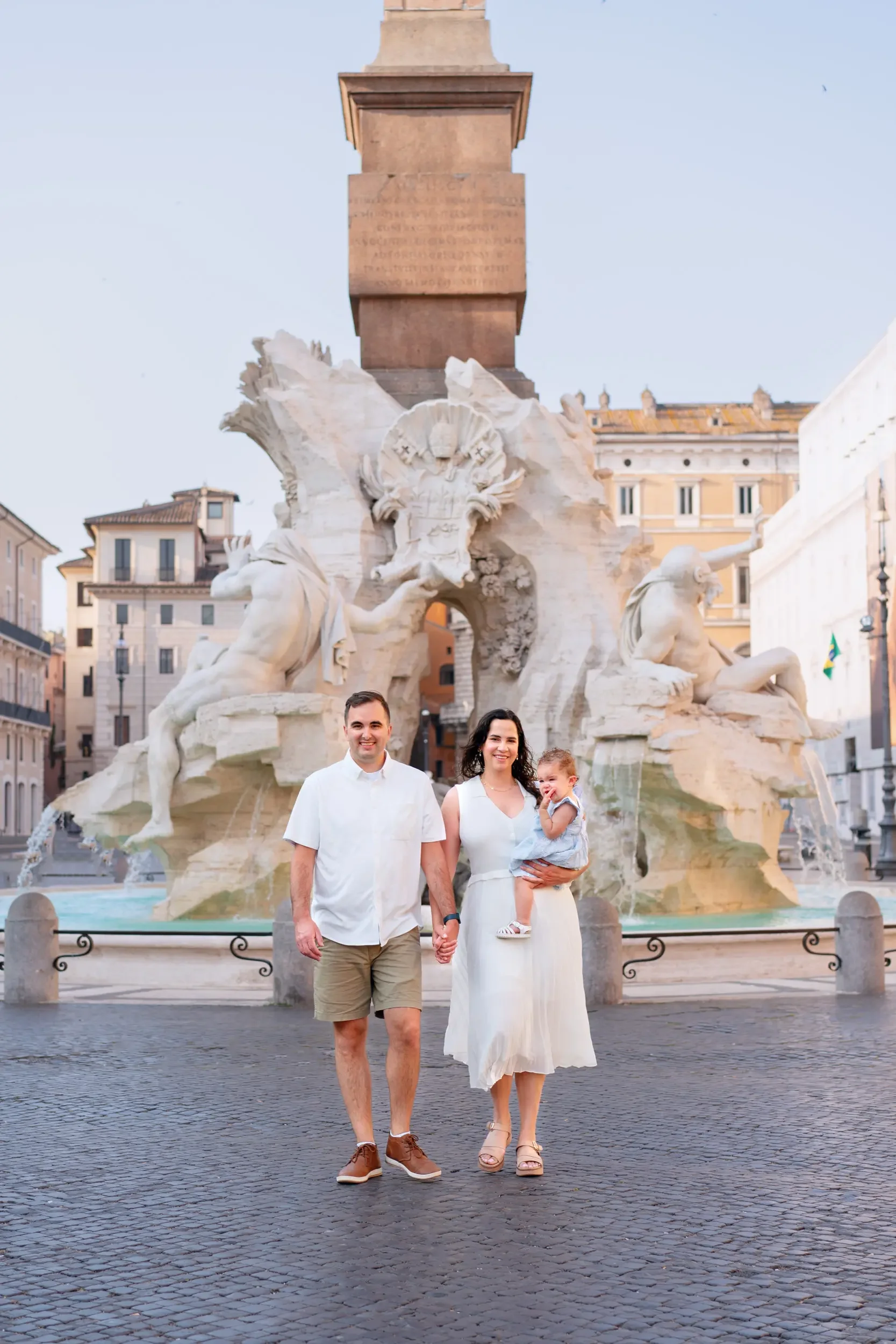 A family of three holding hands in front of the Fountain of the Four Rivers in Rome, Italy, with historic buildings in the background.