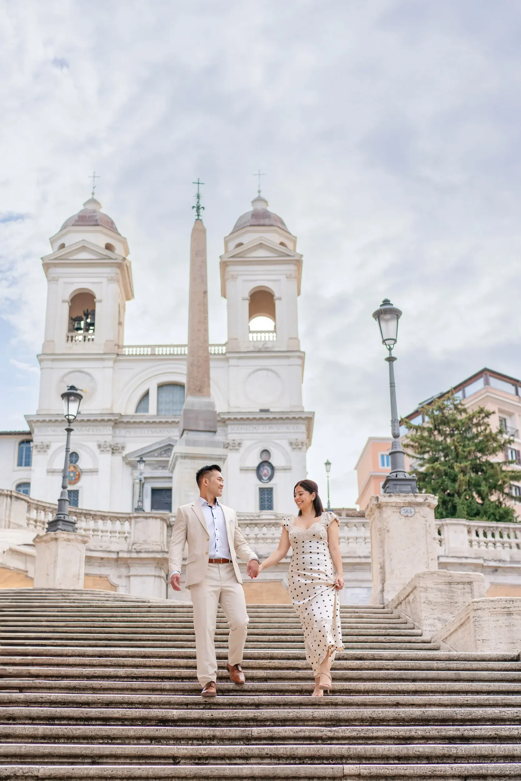 A smiling couple holding hands and walking down stone stairs in front of a large historic church with two bell towers and an obelisk in Rome, Italy.