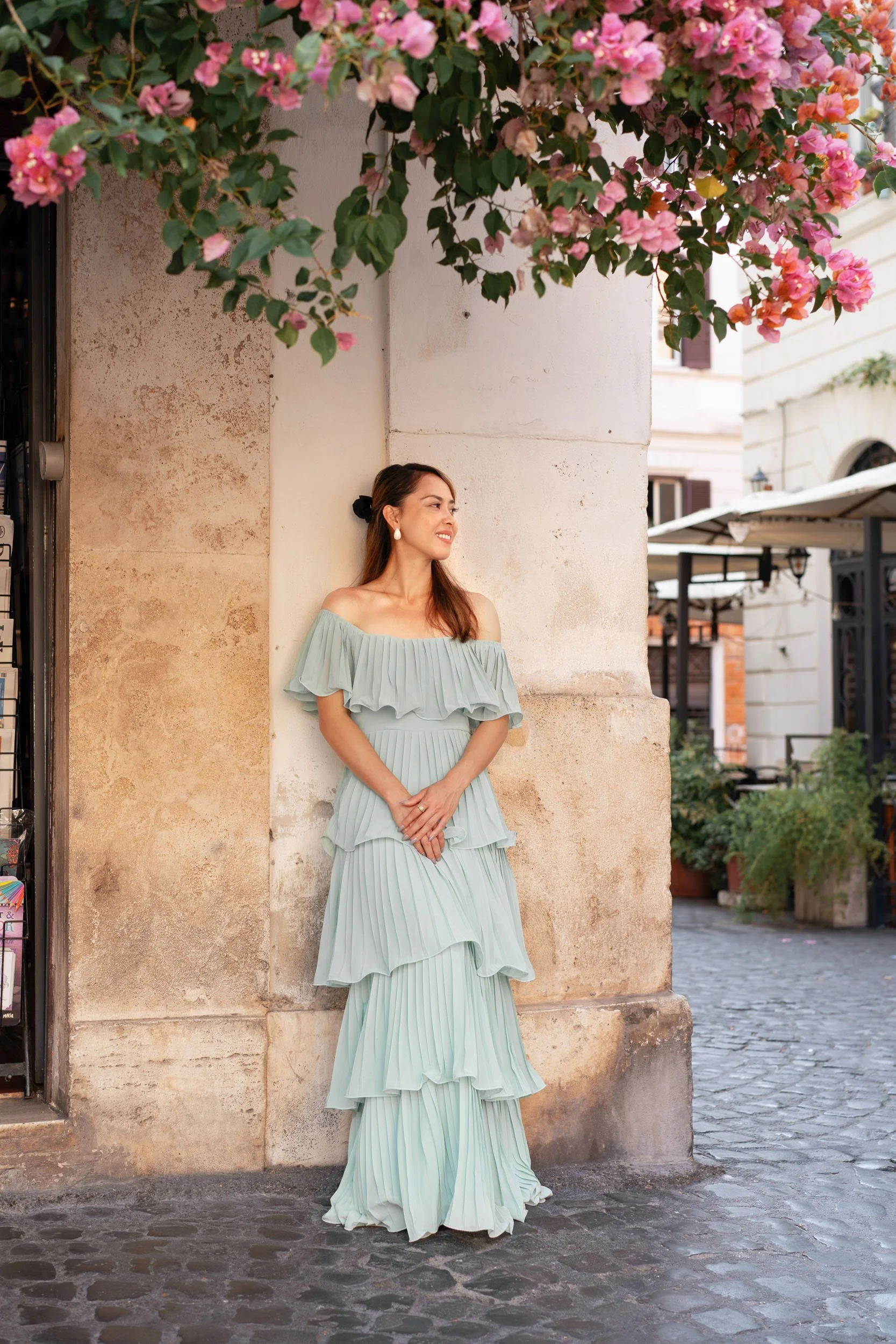 A woman in a light blue, off-shoulder, tiered, pleated dress leaning against a stone column under pink flowering bougainvillea on a cobblestone street.