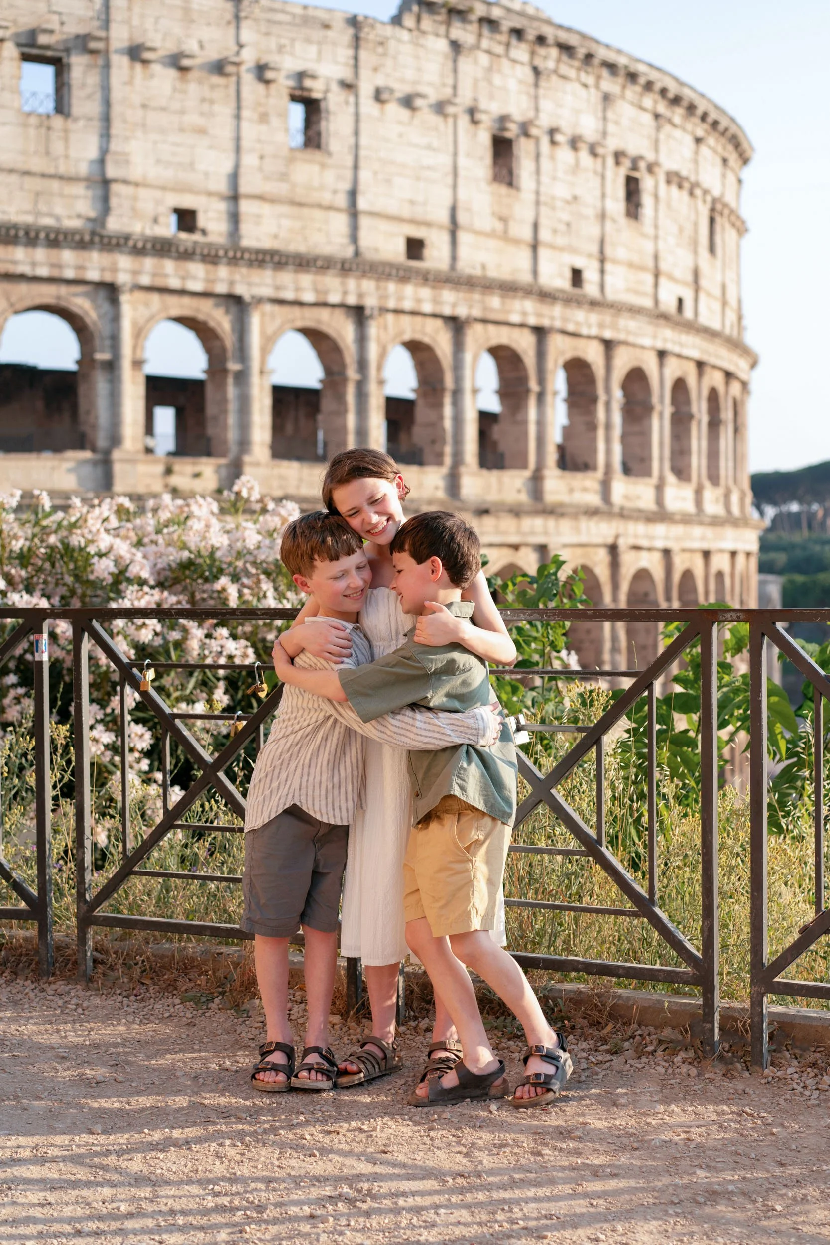 Three children hugging and smiling in front of the Roman Colosseum, with blooming flowers and a metal fence in the background.