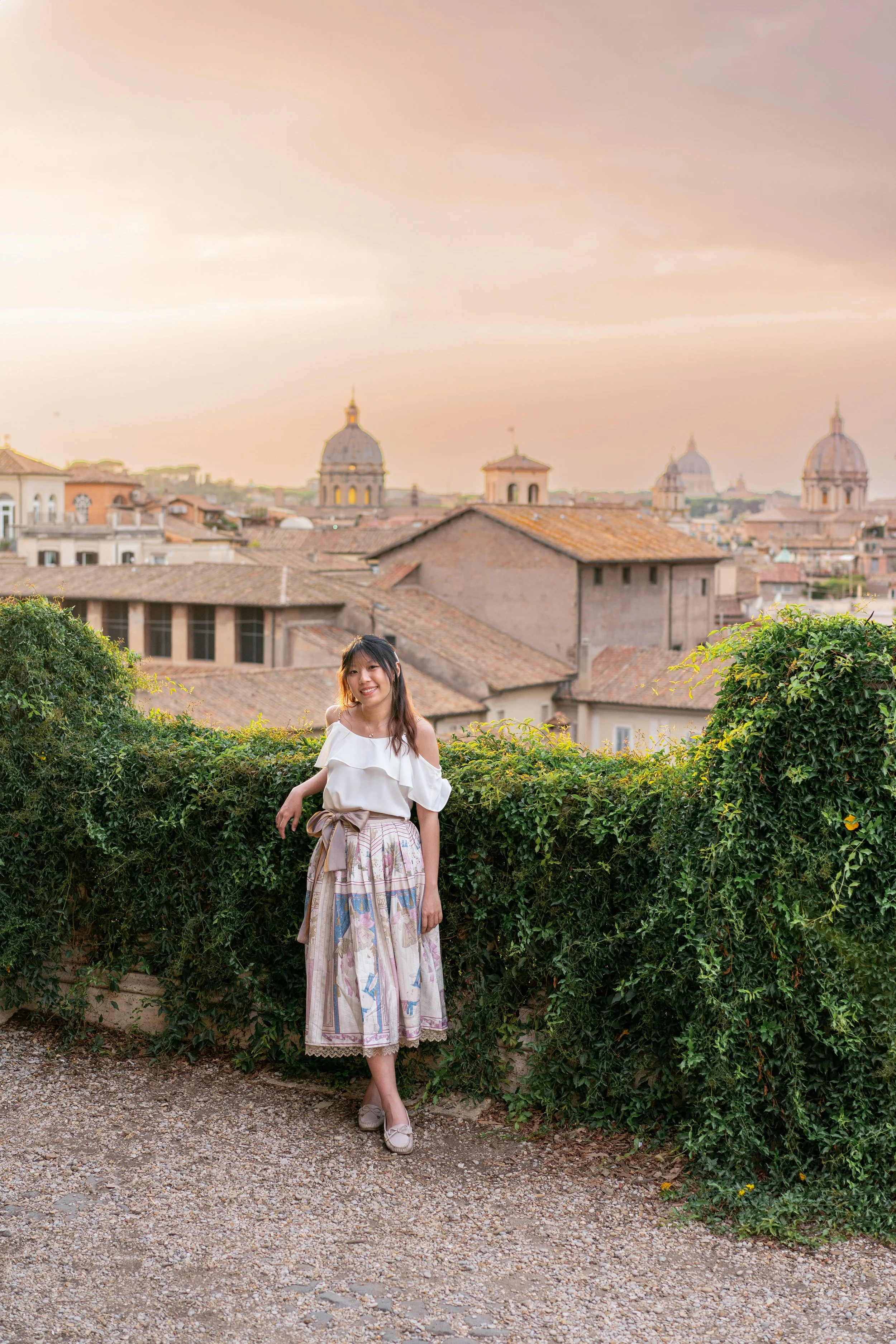 A young woman in a white off-shoulder top and patterned midi skirt standing by a green hedge in front of a cityscape with historic buildings and domed rooftops at sunset.