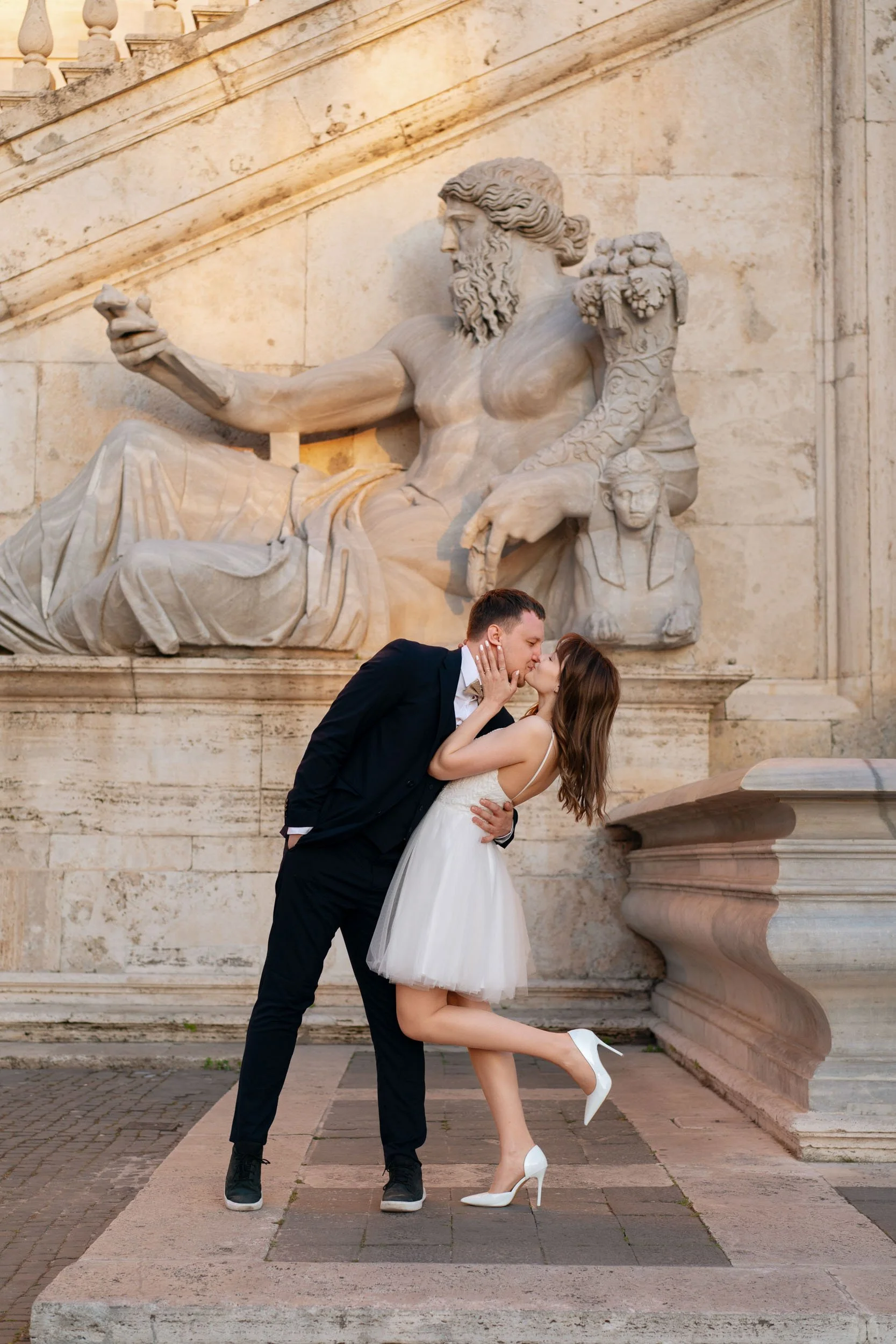 A couple dressed in wedding attire sharing a kiss in front of a large classical sculpture of a seated bearded man with long hair, in an outdoor setting.