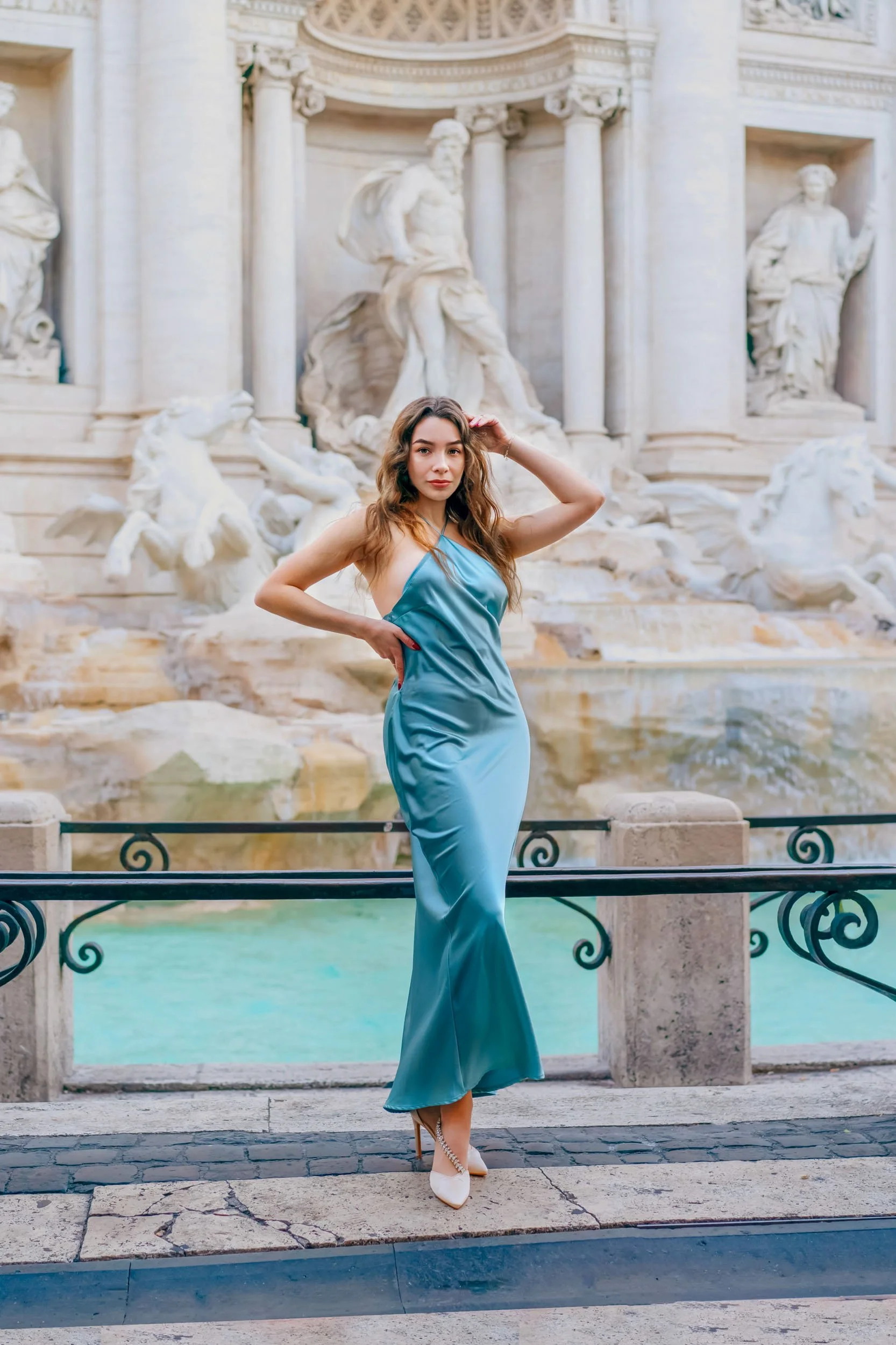 A woman in a shiny blue dress and white heels standing in front of a classical marble fountain with statues.