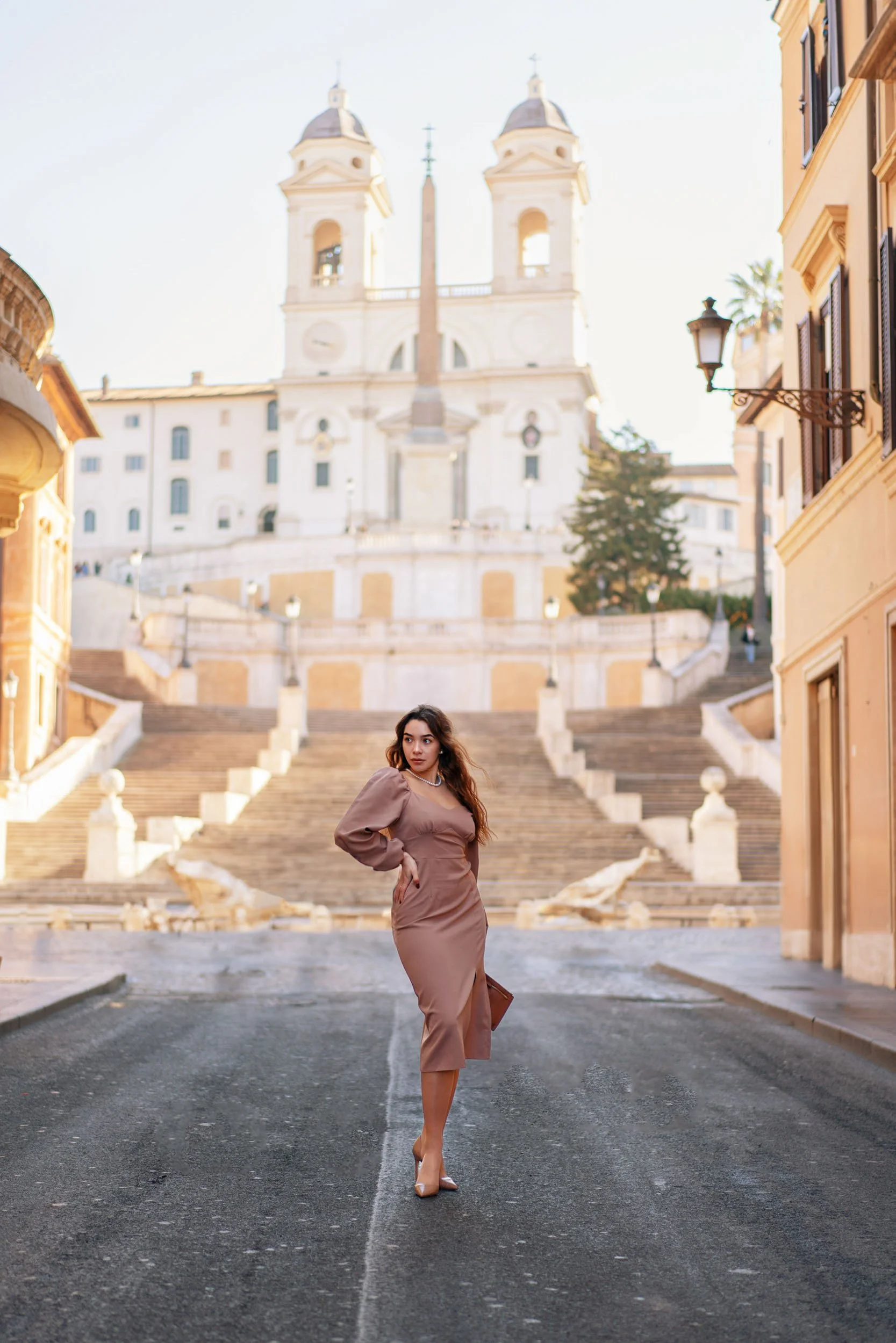 A woman in a brown dress and heels stands on a street with the Spanish Steps and Trinità dei Monti church in Rome in the background.