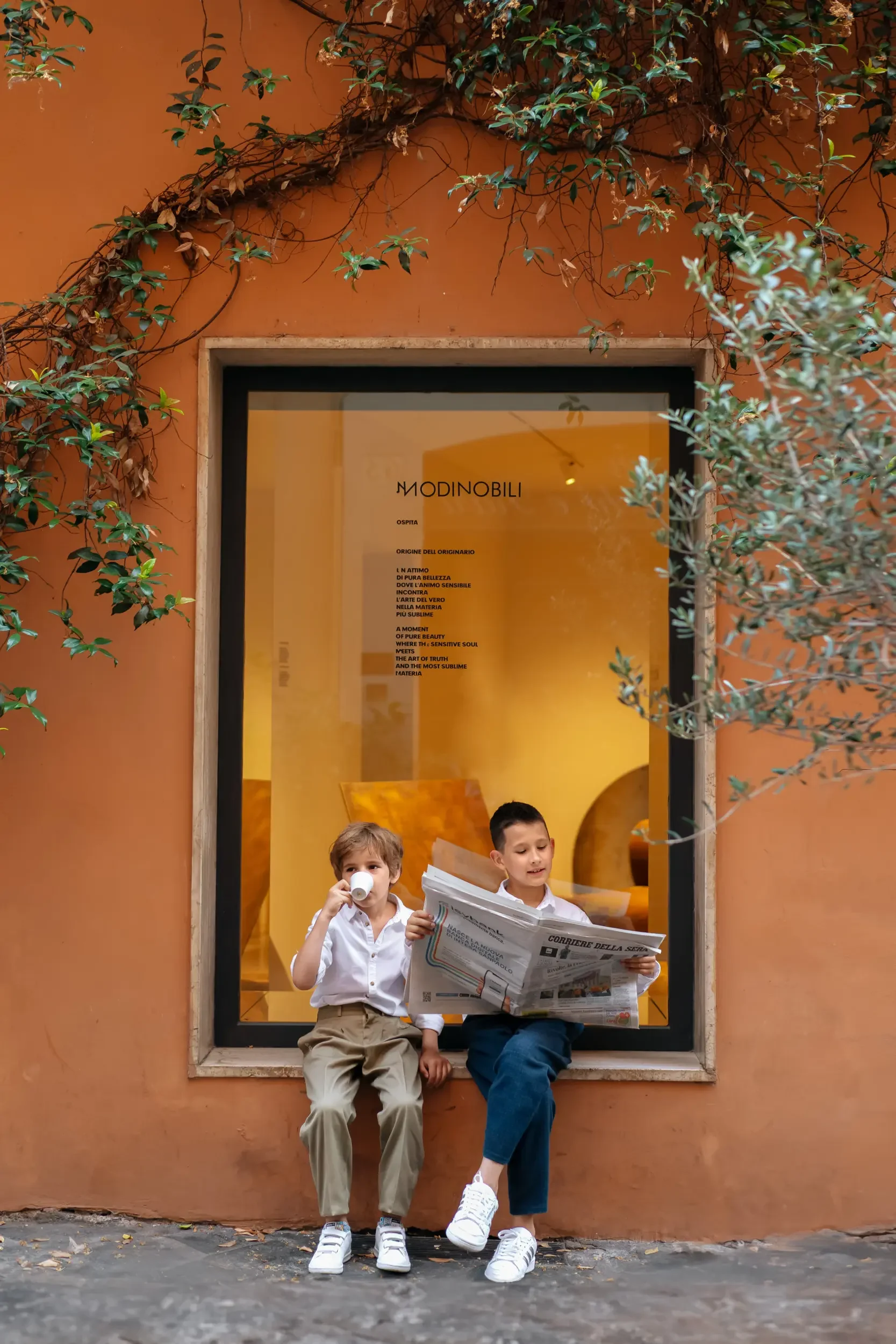 Two young boys sit on the ledge outside a building with orange walls. One boy drinks from a cup, while the other reads a newspaper. Behind them, a large window displays a yellow interior with text on the glass.