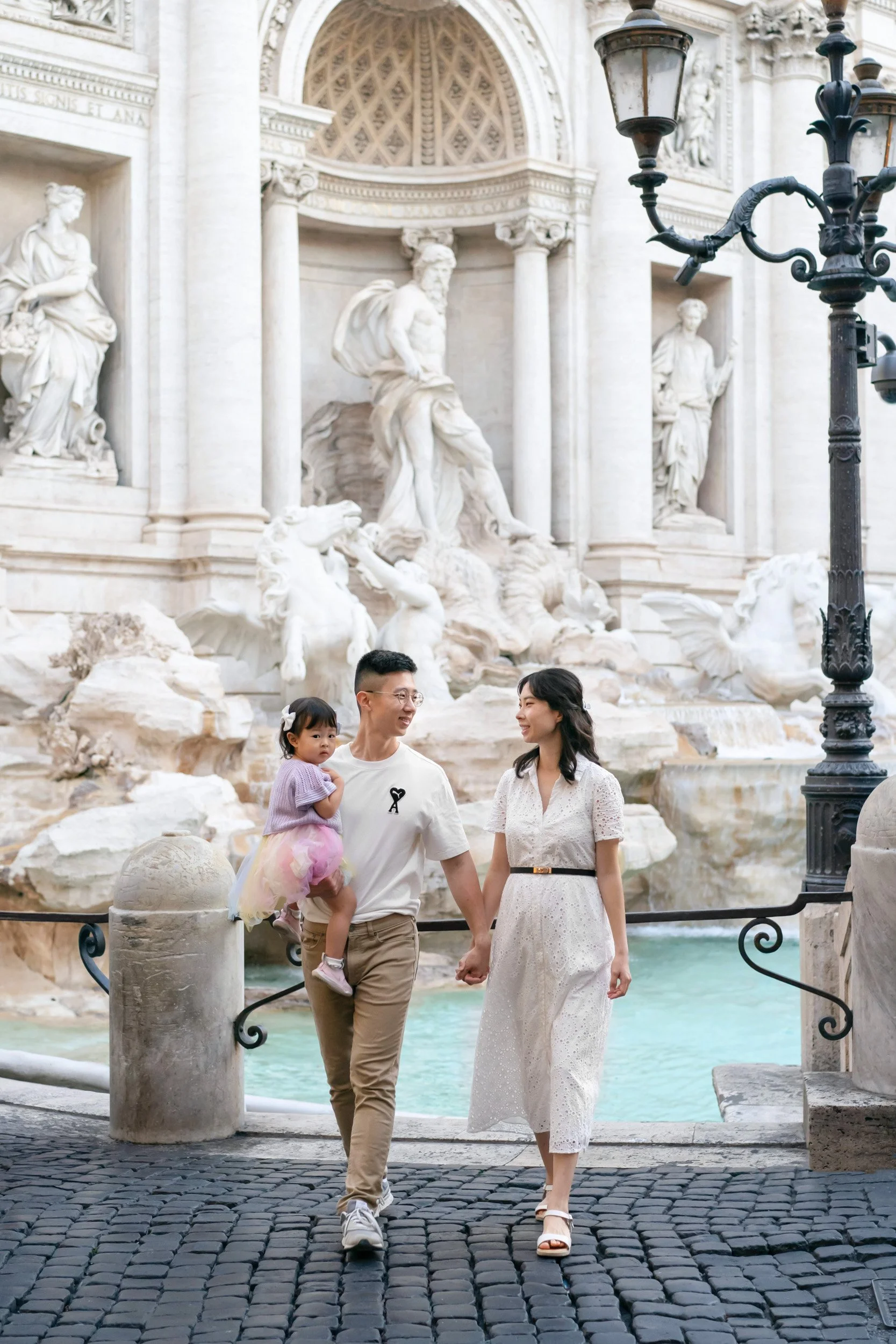 A family of three walking hand-in-hand near the Trevi Fountain in Rome, Italy, with the father carrying a young girl. The fountain features elaborate white marble sculptures and statues.