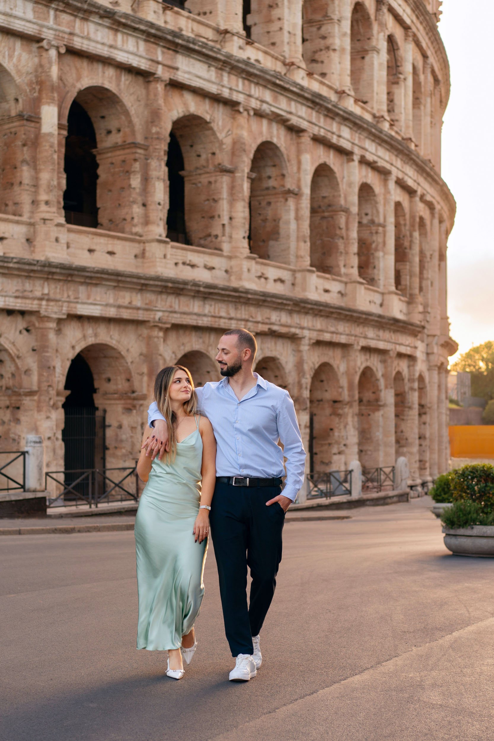 A couple walking together on a street near the Colosseum in Rome at sunset, with the man putting his arm around the woman's shoulders. The woman is wearing a light green satin dress and white heels, and the man is dressed in a light blue shirt and da