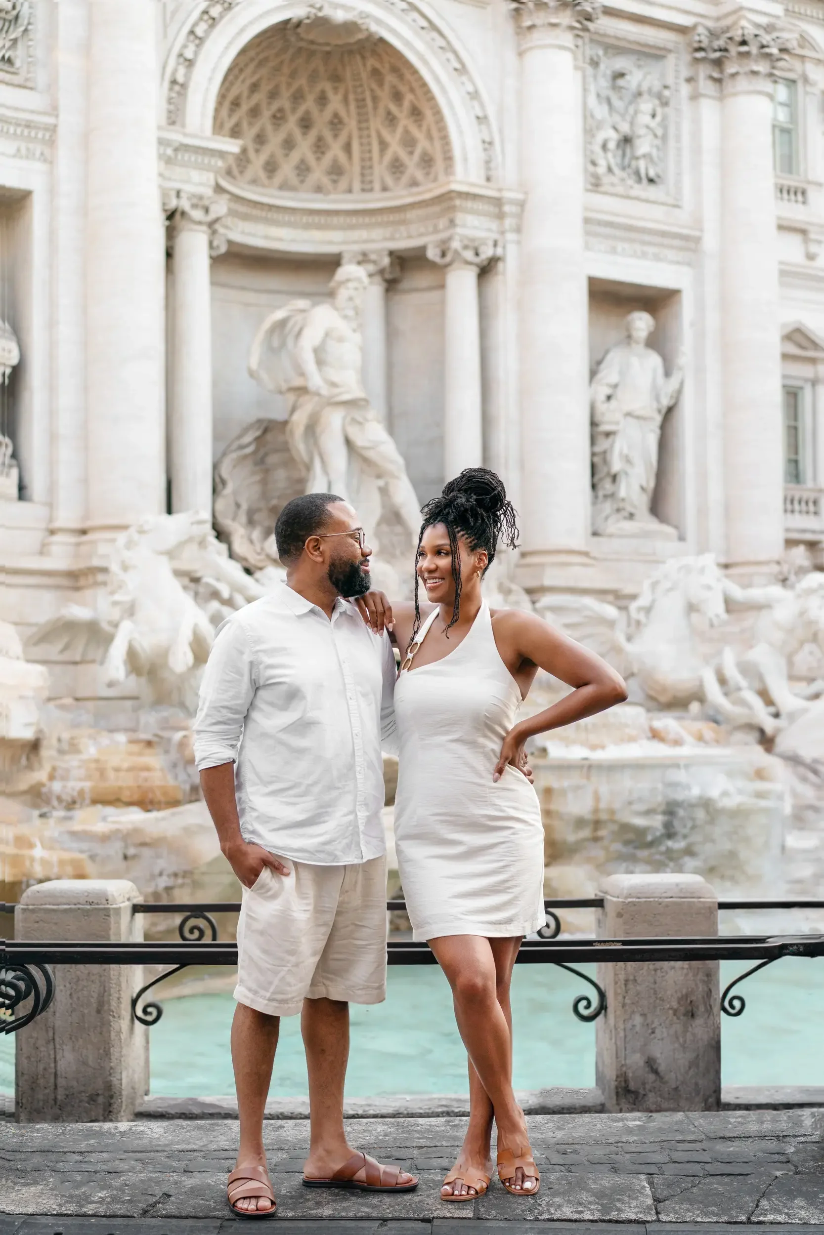 A man and woman in light-colored clothing standing in front of a classical fountain with statues and ornate architecture, smiling and looking at each other.