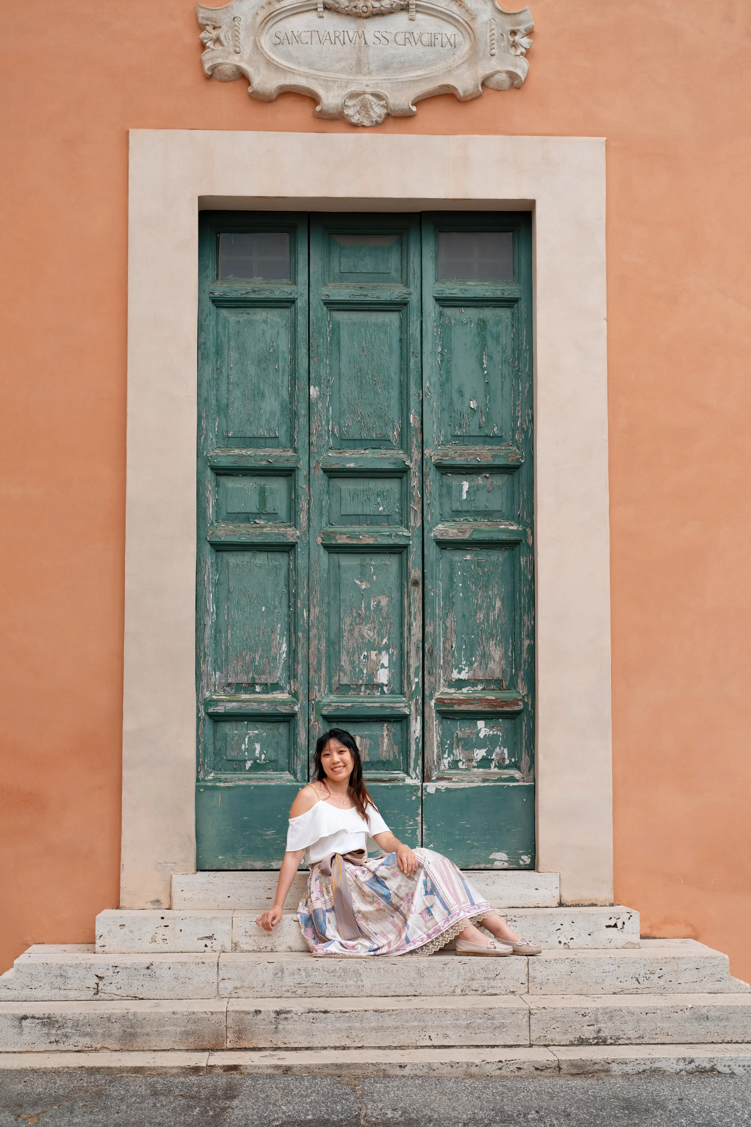 A smiling young woman sitting on steps in front of a large, weathered green wooden door with peeling paint, framed by peach-colored walls and an ornate stone plaque above with Latin inscription.