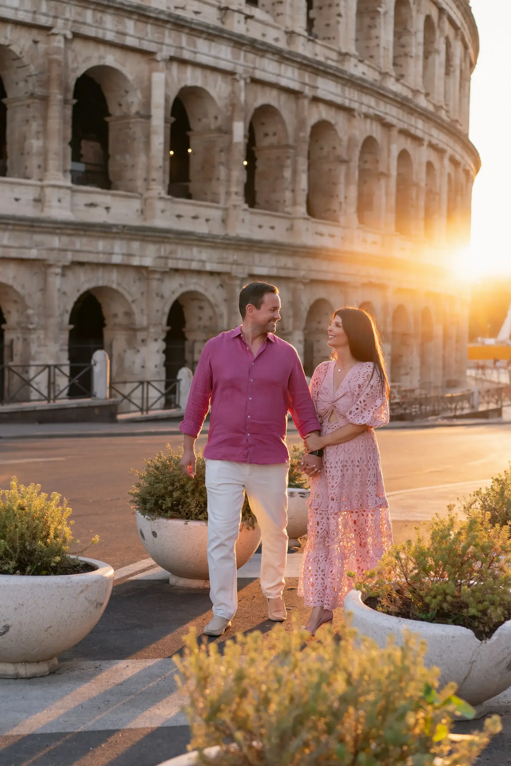 A couple walking hand in hand near the Colosseum at sunset in Rome, Italy, with warm sunlight illuminating the scene and potted plants on the pavement.
