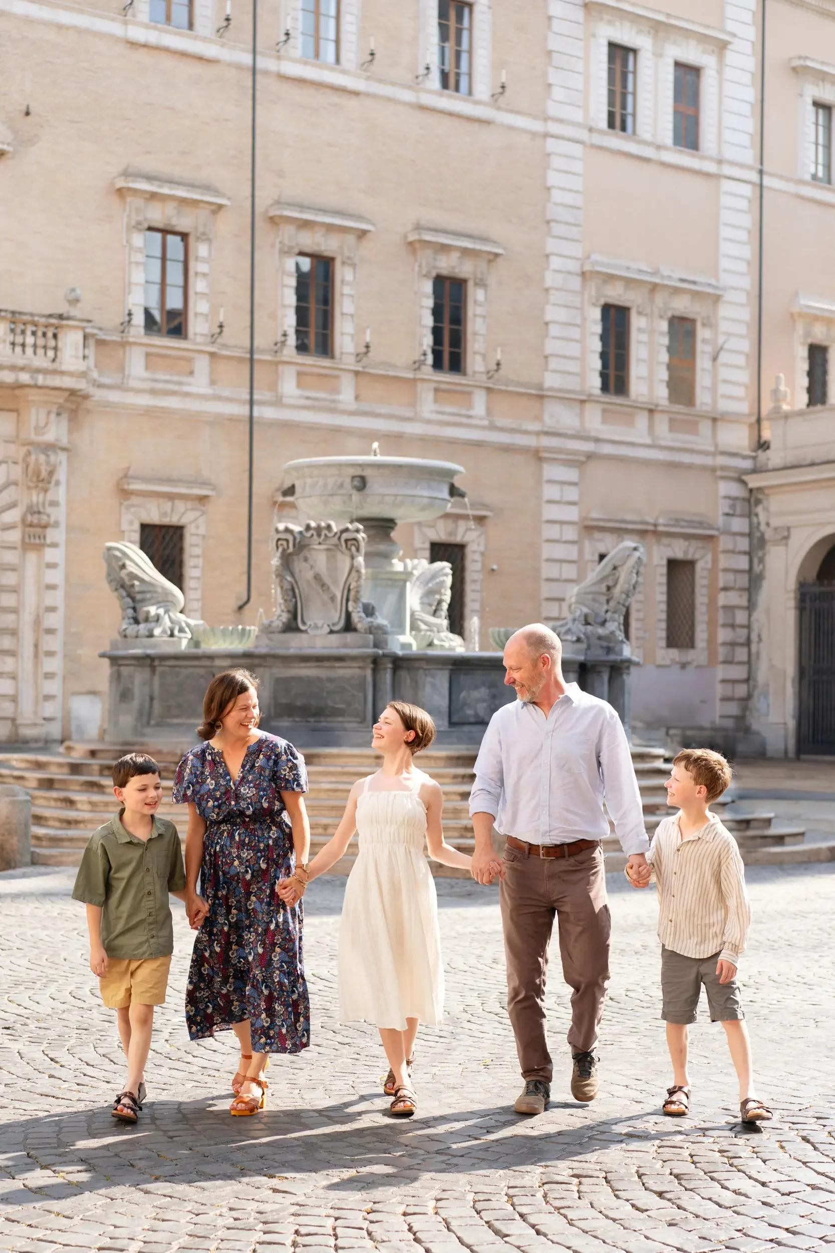 A family of six holding hands and walking together on a cobblestone street in front of a historic building with a fountain in the background.