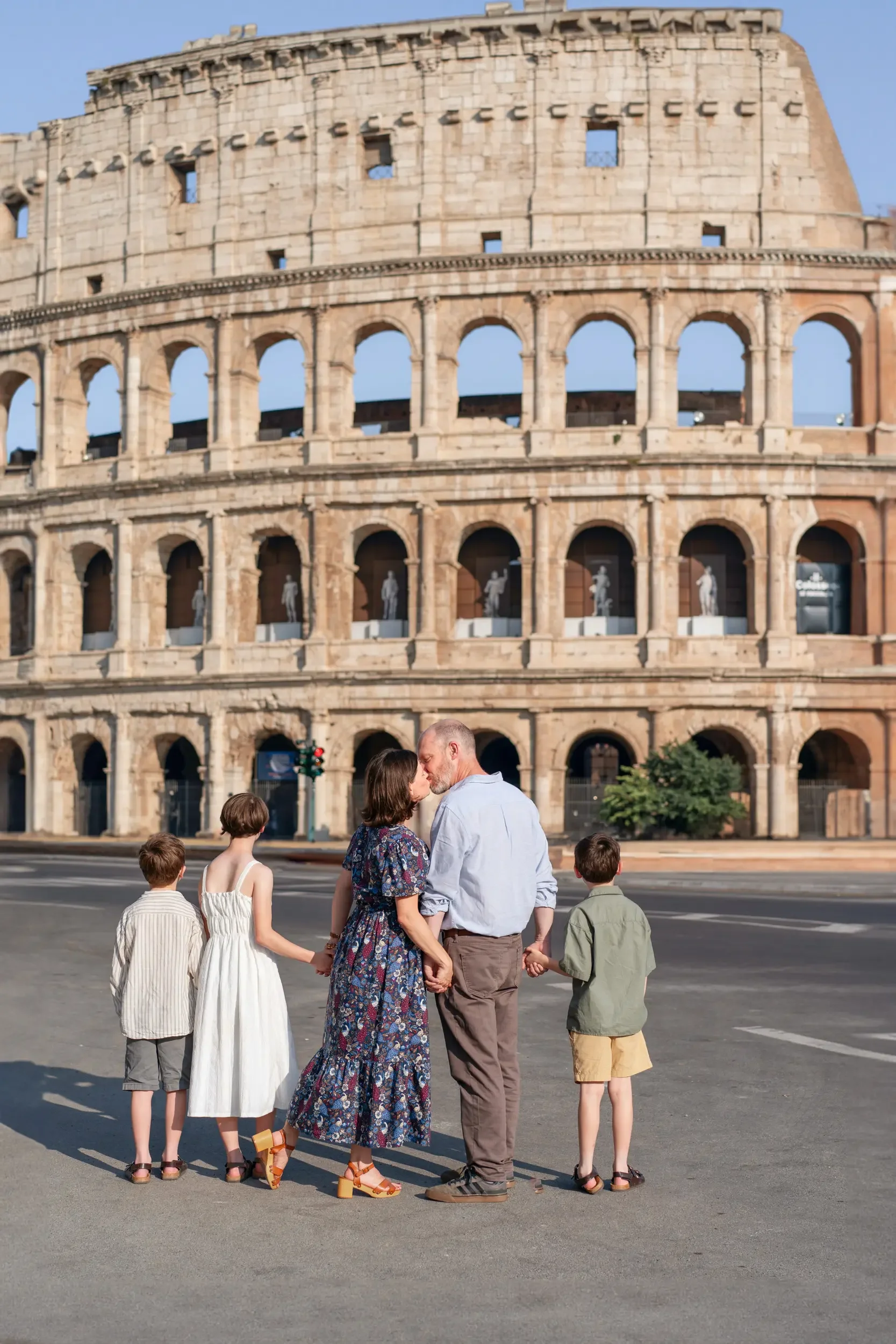 A couple kissing and holding hands, surrounded by children, standing on a street in front of the Roman Colosseum in Rome, Italy.