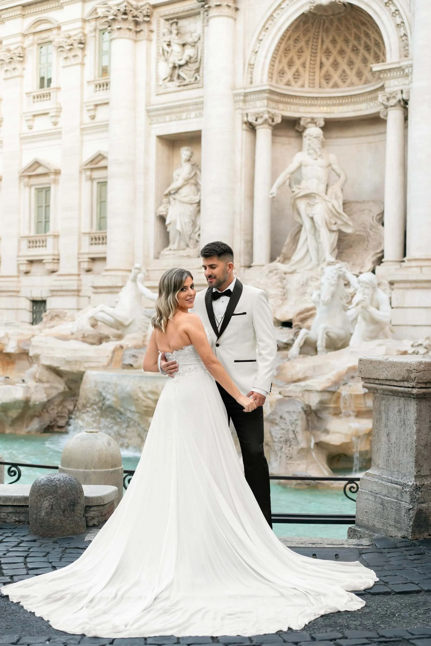 A newlywed couple dressed in wedding attire, standing in front of the Trevi Fountain in Rome, Italy.