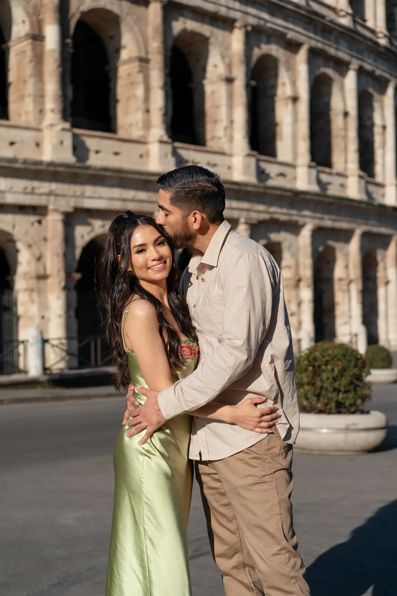 A couple standing close together in front of the Colosseum in Rome, Italy. The woman is smiling and wearing a light green dress, while the man is kissing her on the forehead and wearing a beige shirt with khaki pants.