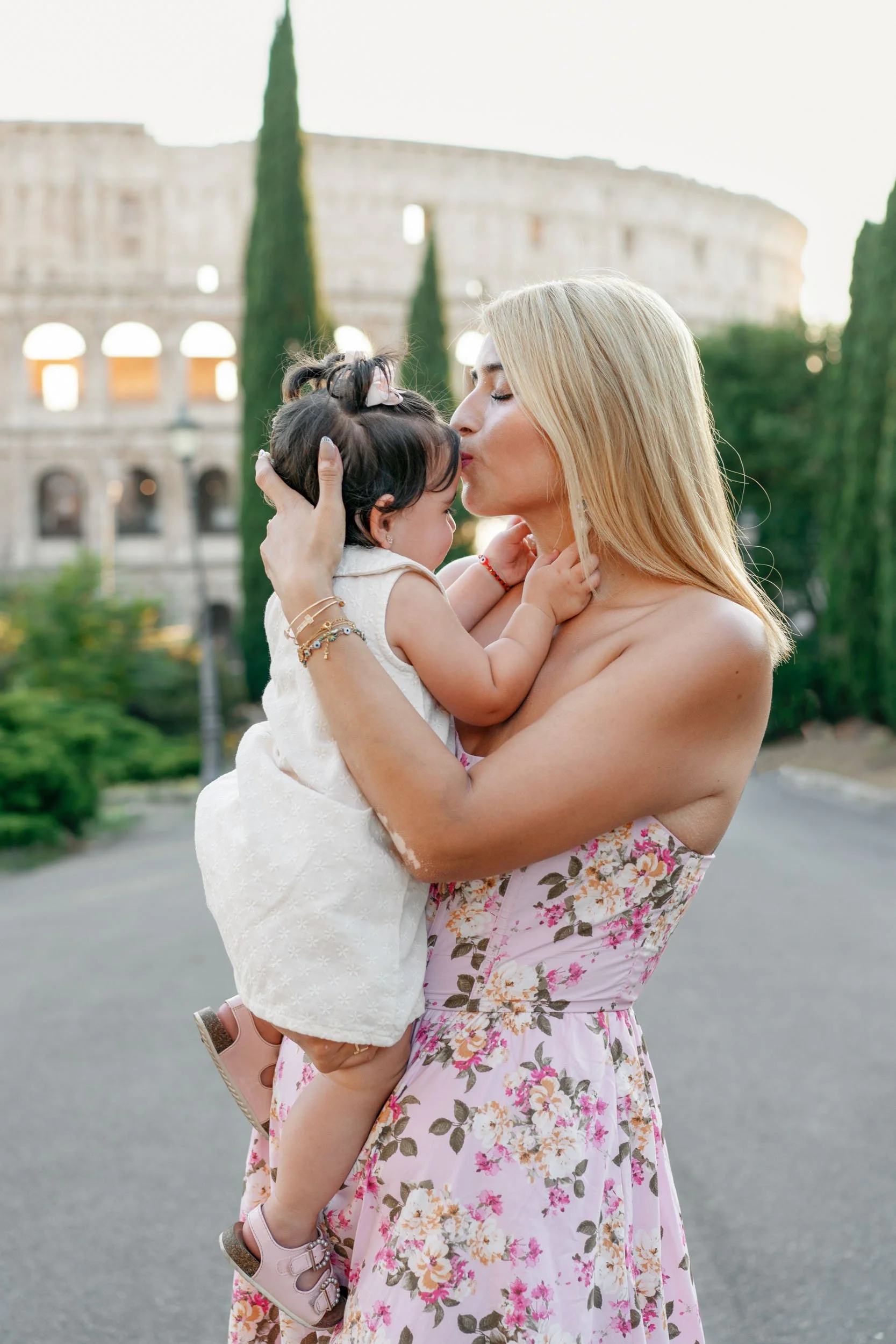 A woman with blonde hair wearing a pink floral dress holds and kisses a young girl with dark hair in a white dress, with the Colosseum in Rome in the background.