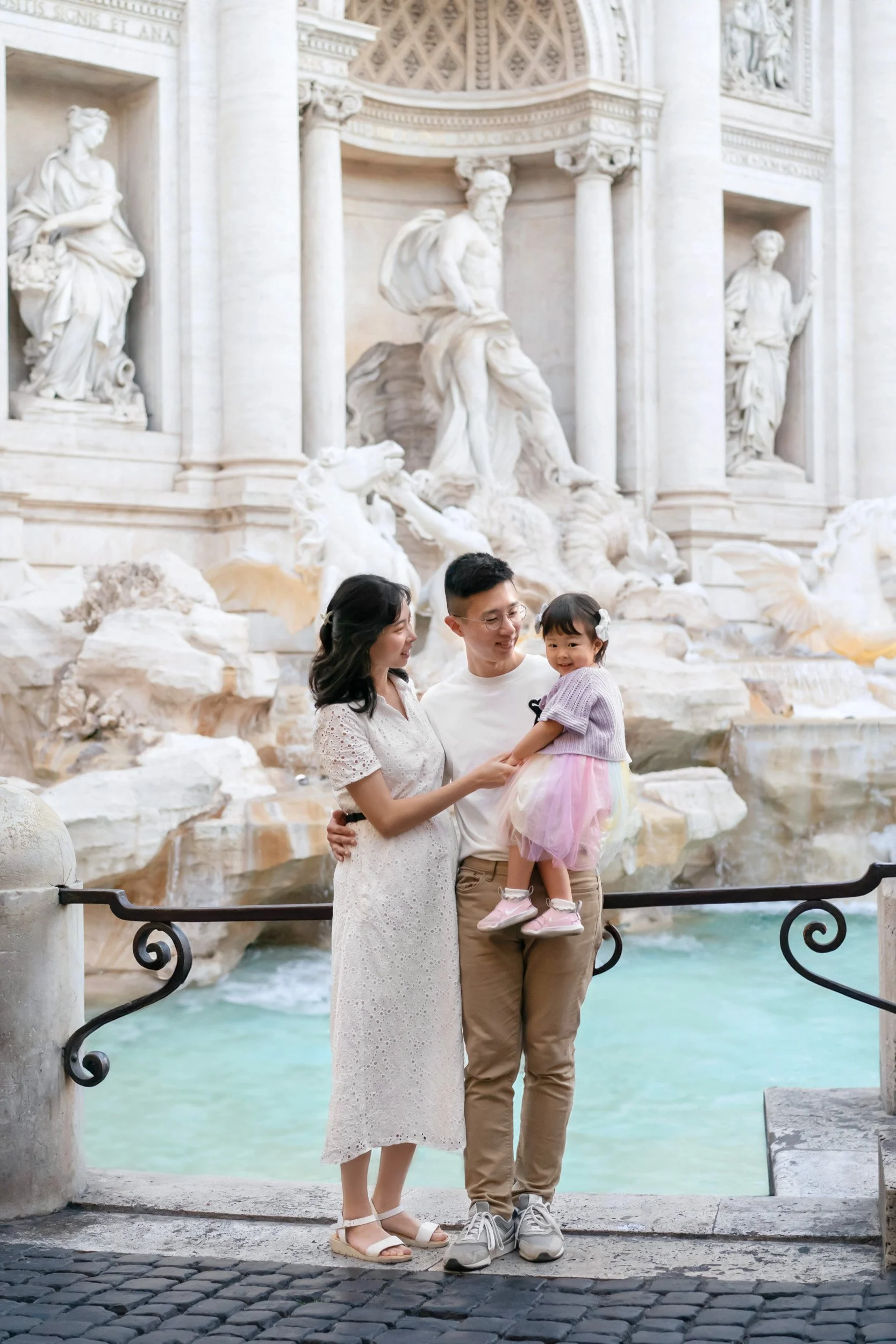 A family of three, including a woman, a man, and a young girl, standing in front of the Trevi Fountain in Rome, Italy. The little girl is being held by the man, and they are all smiling.