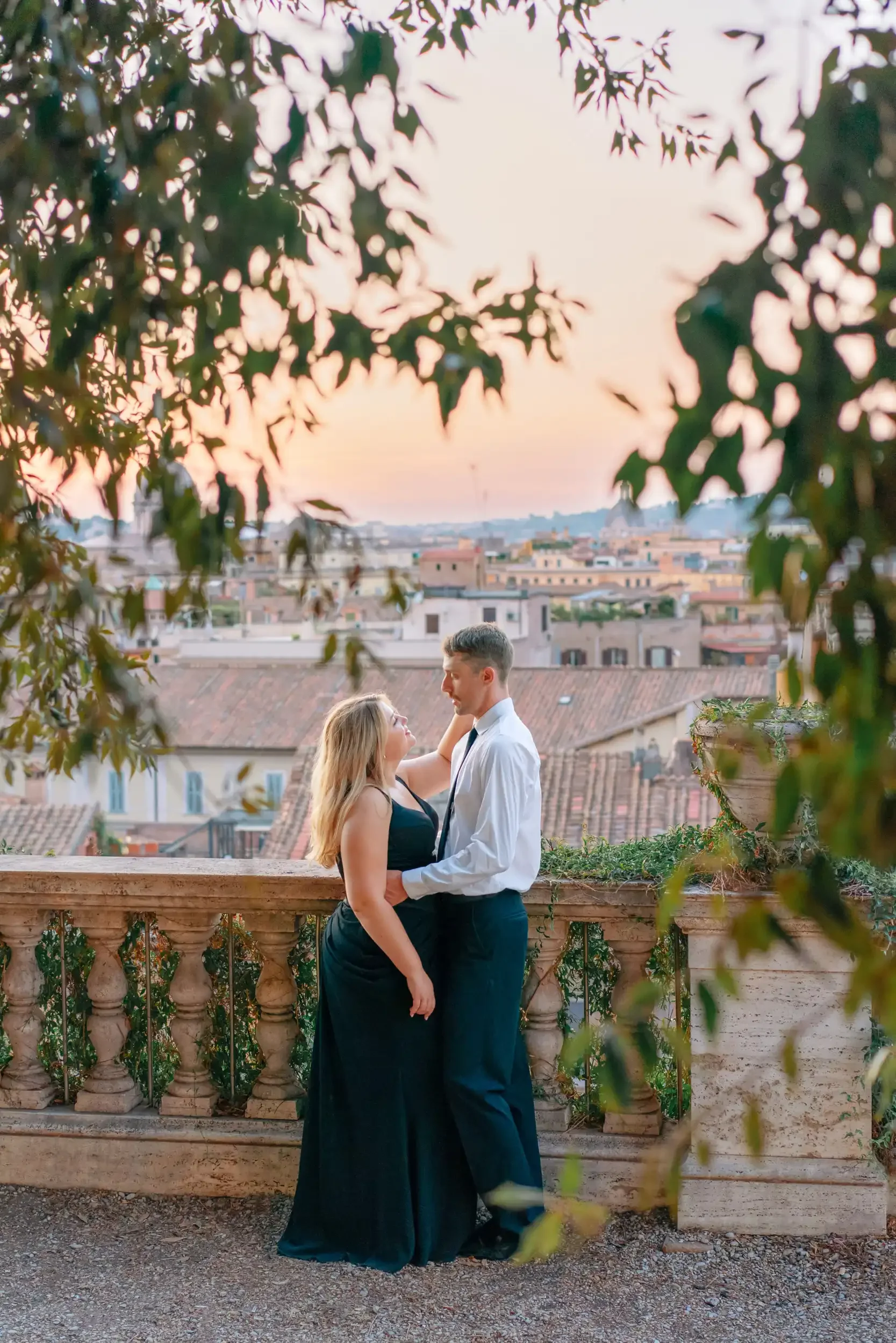 A couple standing close together on a balcony overlooking a cityscape at sunset, surrounded by foliage.