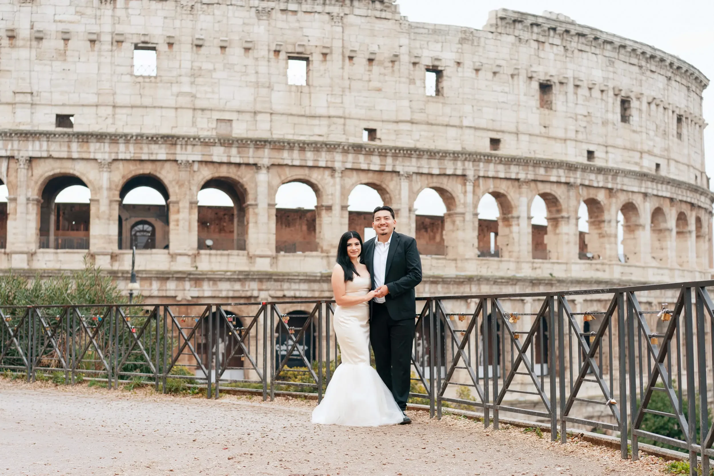 A couple dressed in formal wedding attire standing in front of the Colosseum in Rome, Italy.