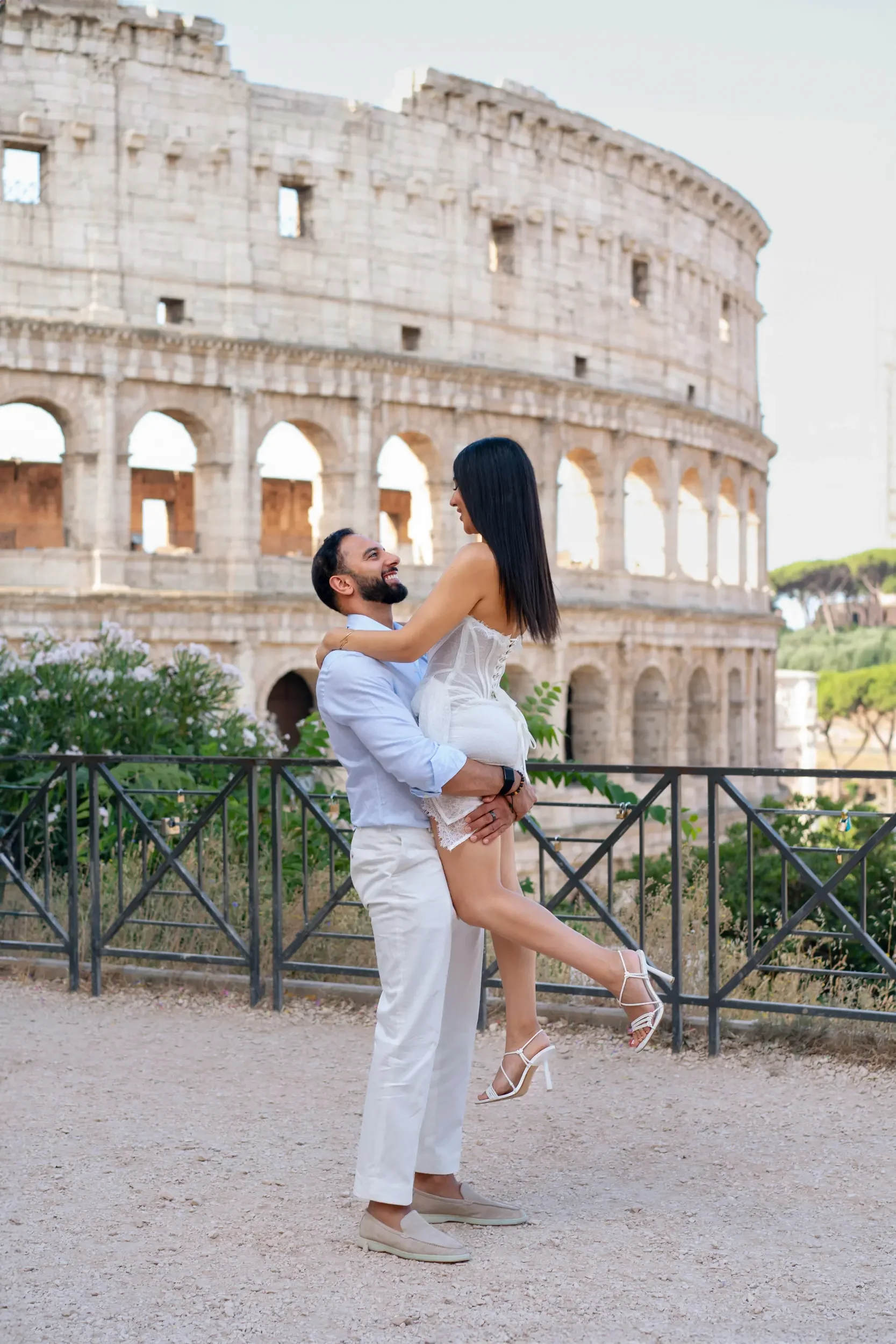 A happy couple in white clothing, with the man lifting the woman, standing in front of the Colosseum in Rome, Italy, during the daytime.