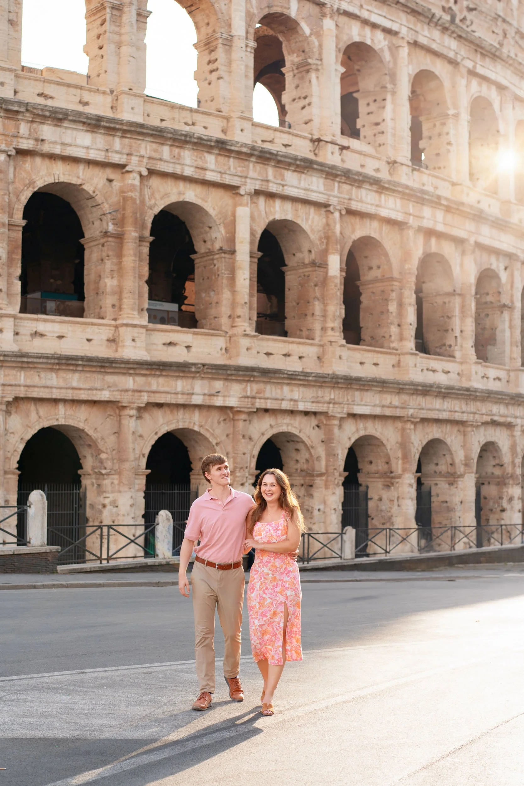 A young couple, a man in a pink shirt and beige pants and a woman in a pink floral dress, walks arm in arm in front of the Colosseum in Rome during sunset.