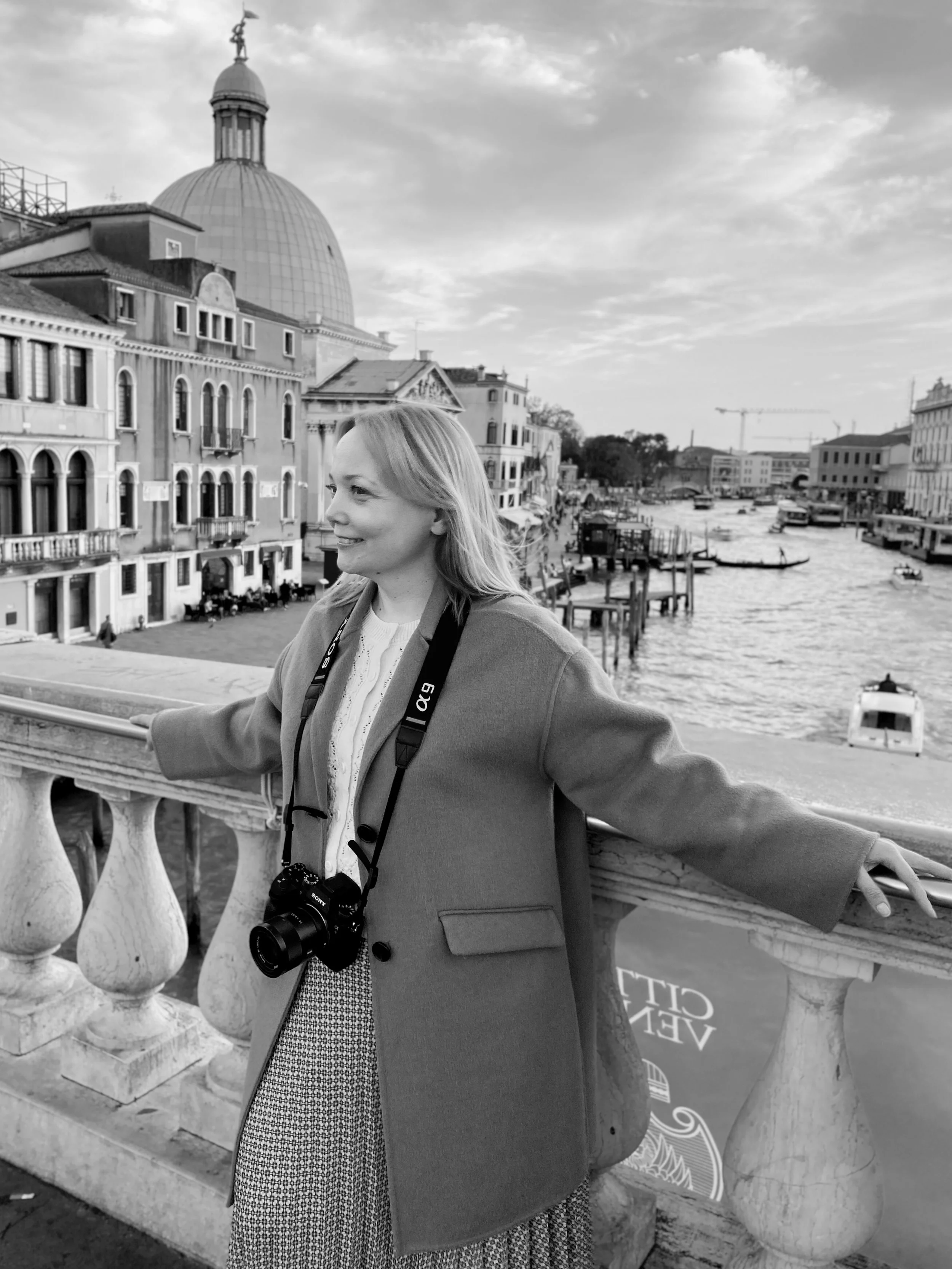 A woman with a camera around her neck standing on a bridge overlooking a canal with historic buildings and boats in Venice, Italy.