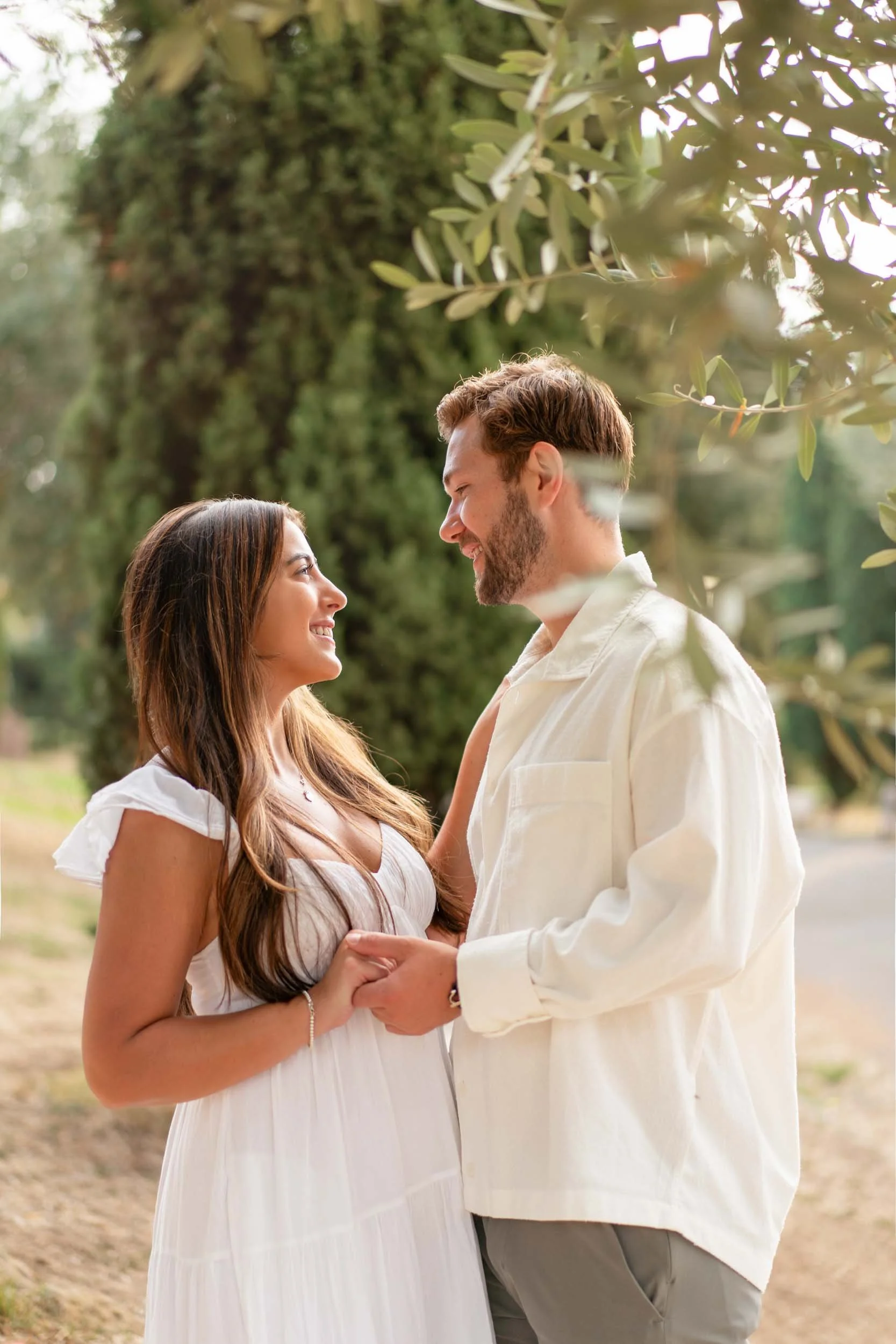 A couple standing outdoors, smiling and holding hands, surrounded by trees and greenery.