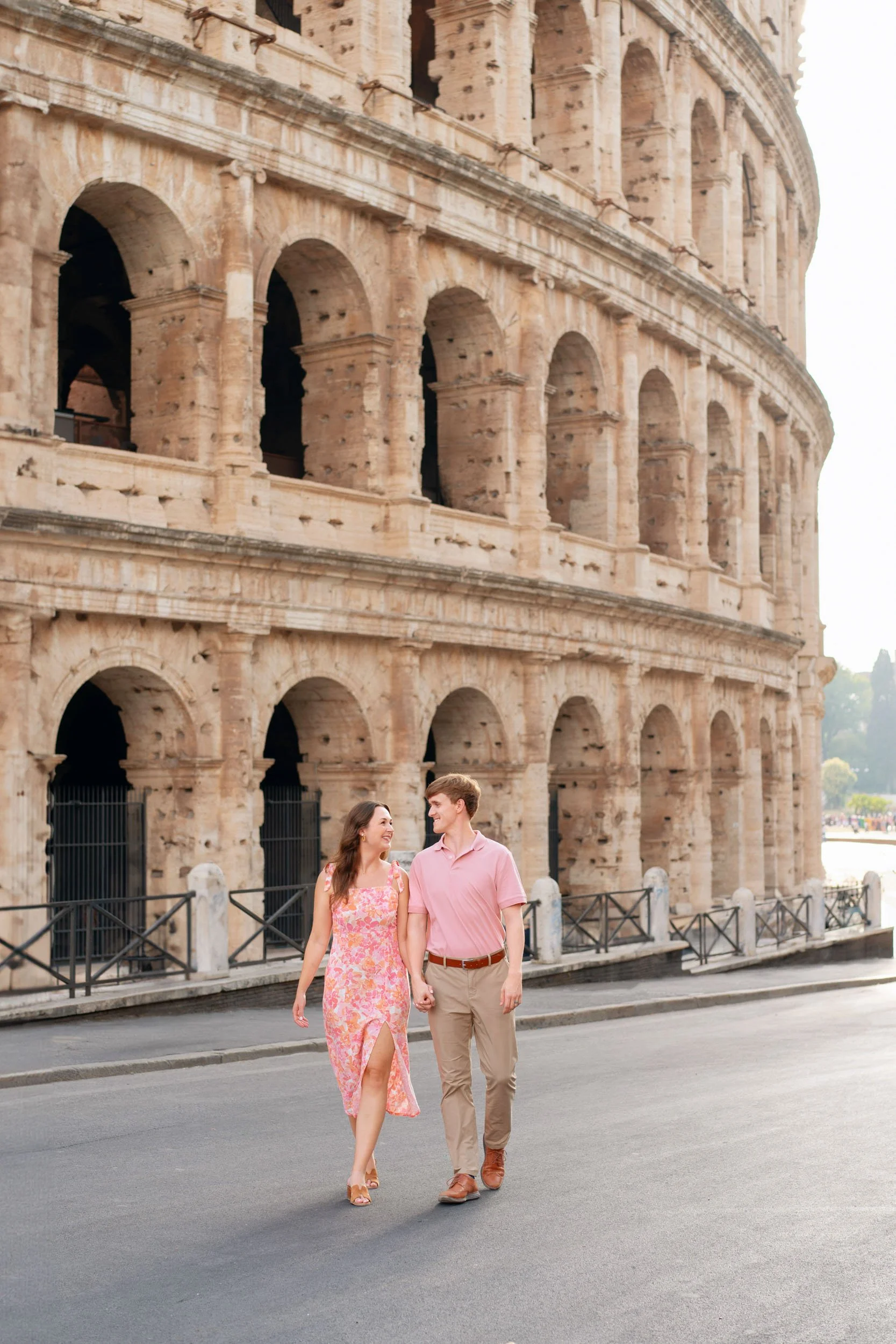 A young couple walking hand-in-hand in front of the Colosseum in Rome, Italy, smiling at each other on a sunny day.