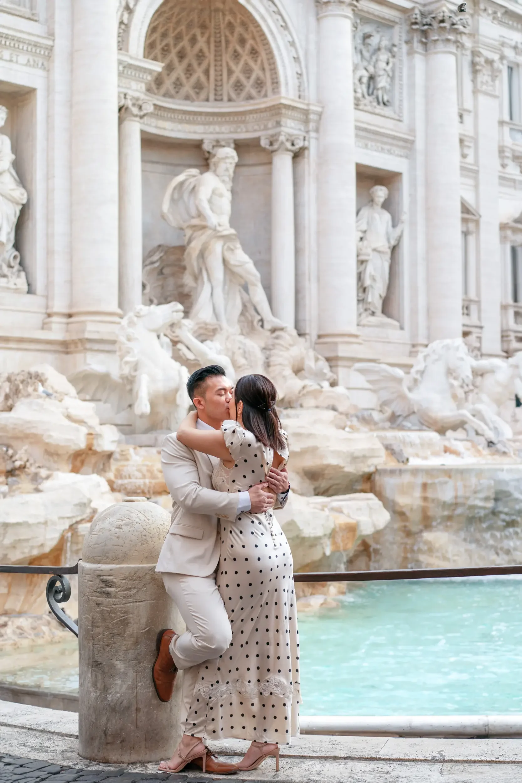 A couple kissing in front of the Trevi Fountain in Rome, Italy. The man is in a light-colored suit and the woman is in a white dress with black polka dots.