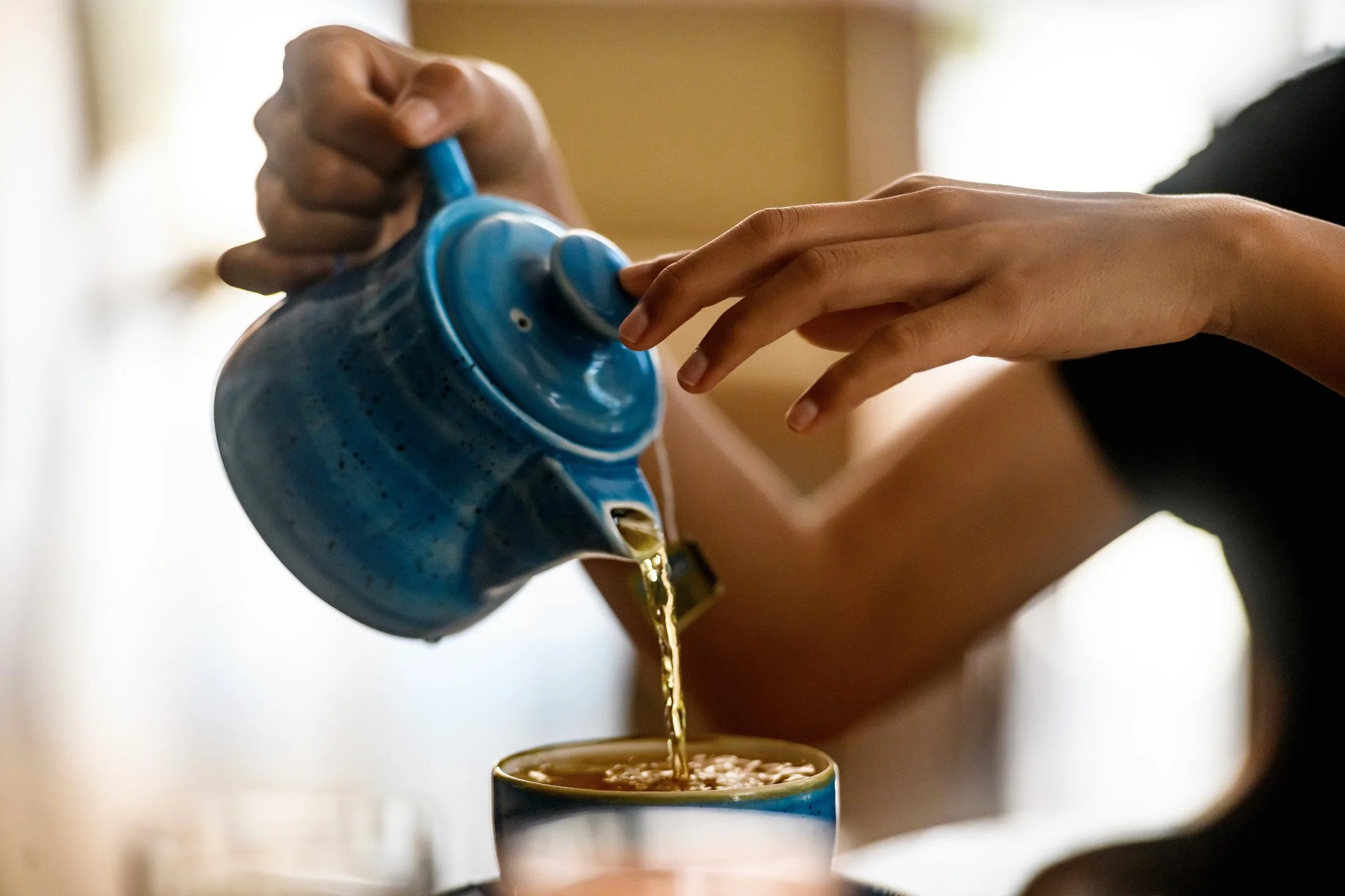 A person pouring a hot beverage, likely coffee or tea, from a blue teapot into a mug.