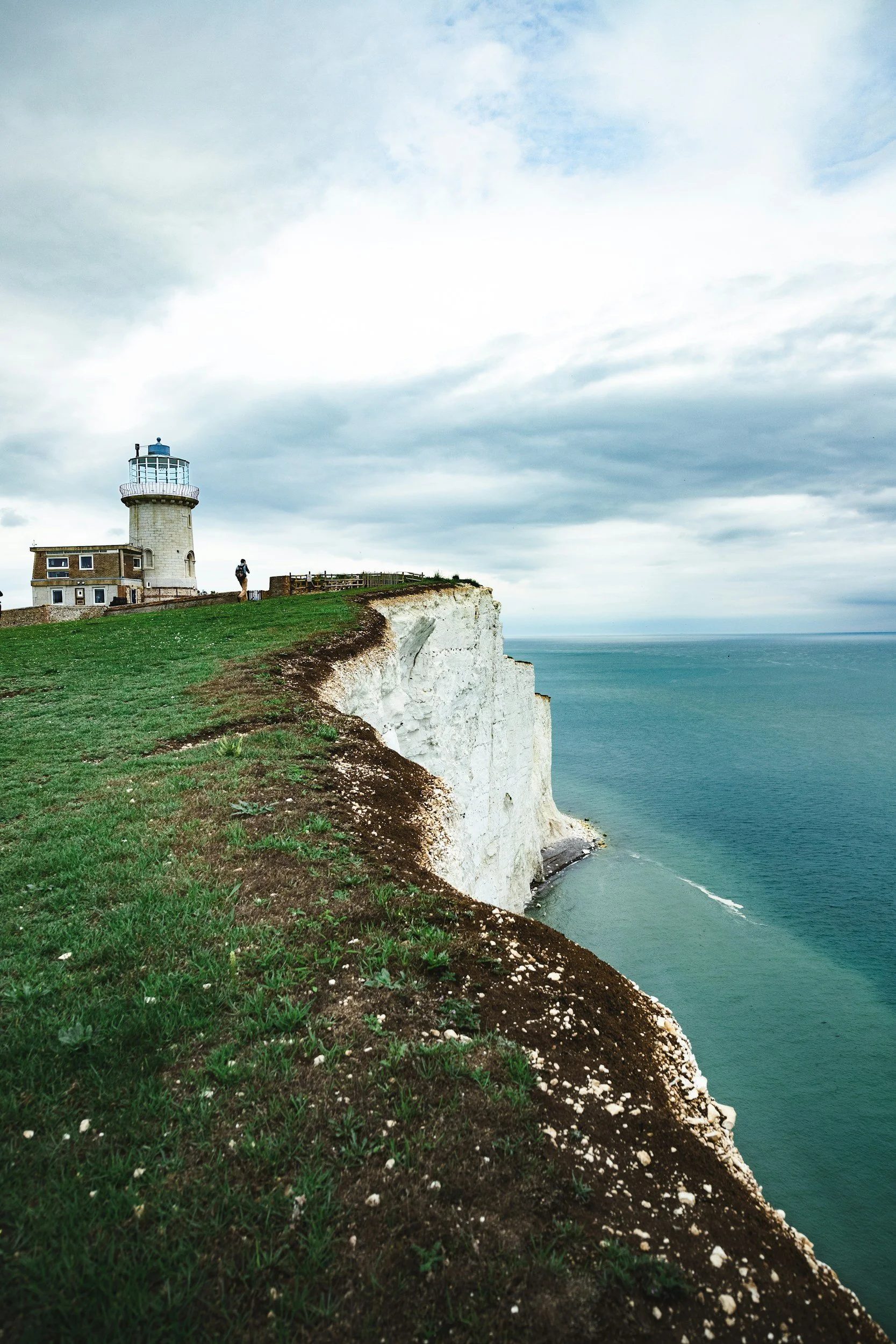 Cliffs with a lighthouse and a person walking on the grass near the edge, overlooking the sea under a cloudy sky.
