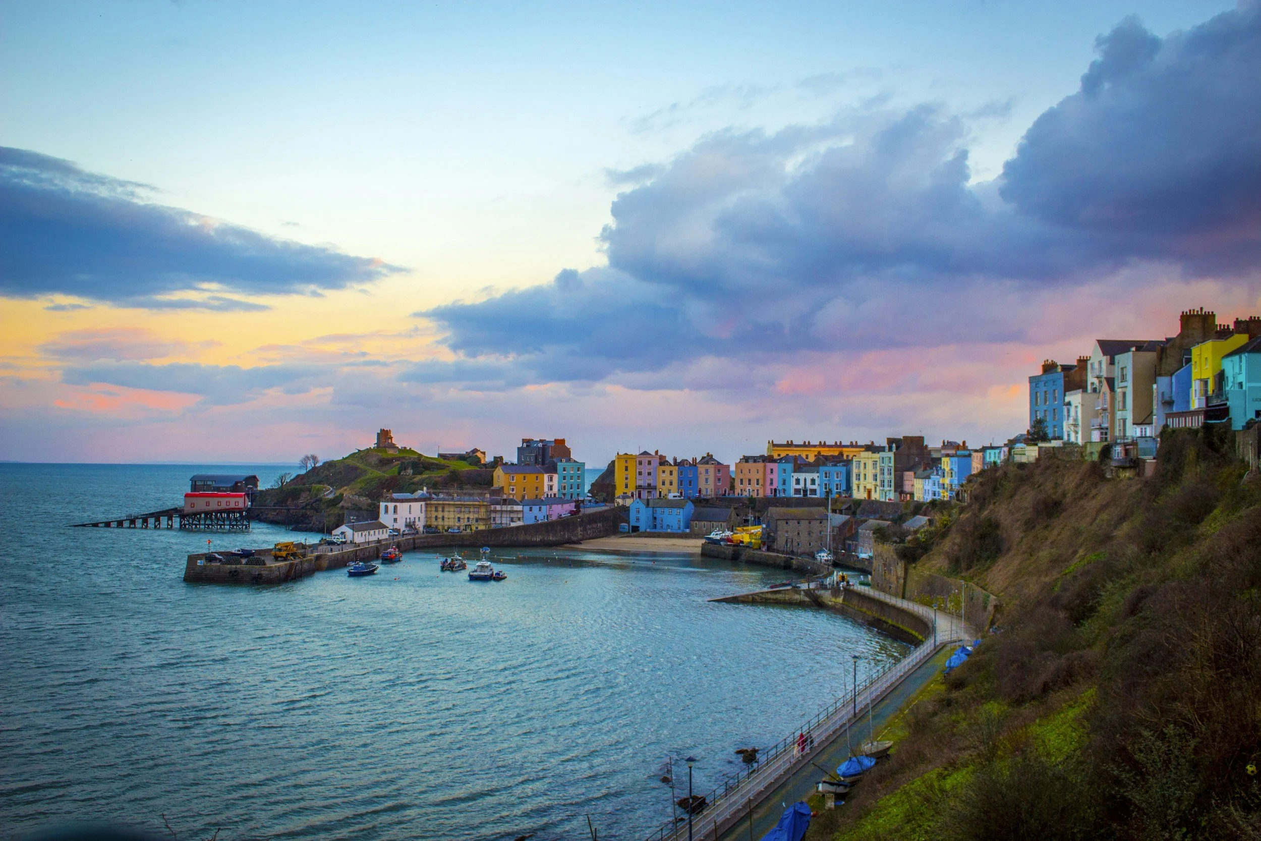 Colorful houses on a hillside by the ocean with a pier and boats, under a sky with clouds at sunset.