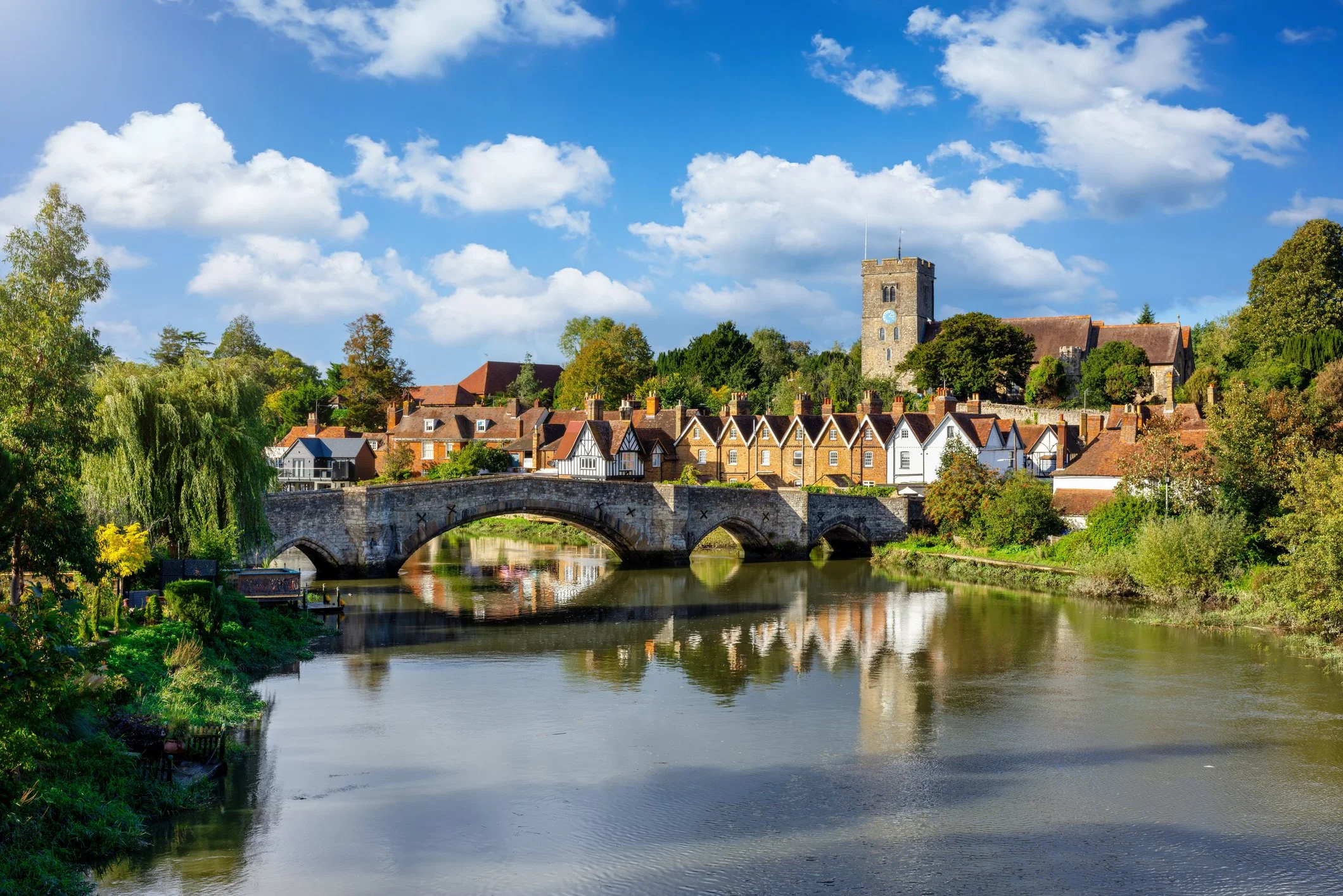 A peaceful riverside town with a stone bridge spanning a calm river, flanked by with lush green trees and quaint houses, including a church with a tower, under a blue sky scattered with fluffy white clouds.