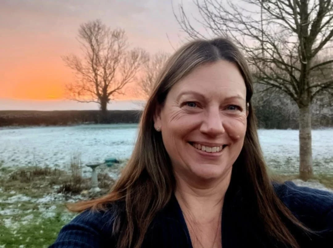 A woman smiling outdoors in a winter landscape at sunset, with snow on the ground and leafless trees in the background.