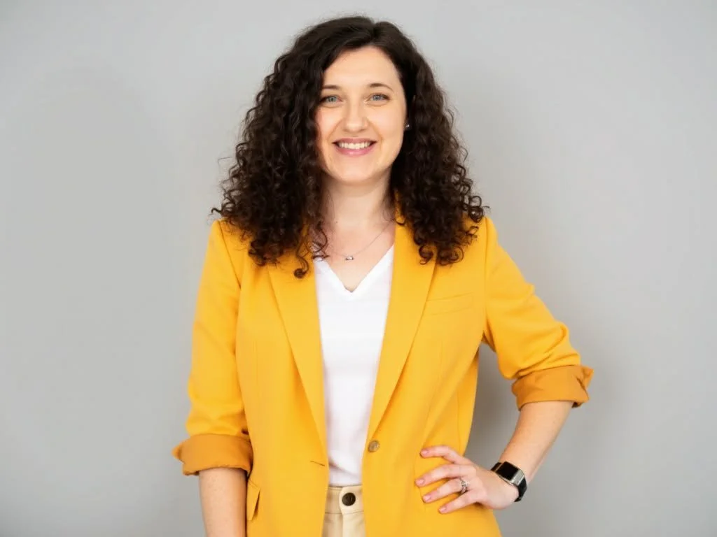 A woman with curly brown hair wearing a yellow blazer, white shirt, and watch, standing against a plain light gray background.