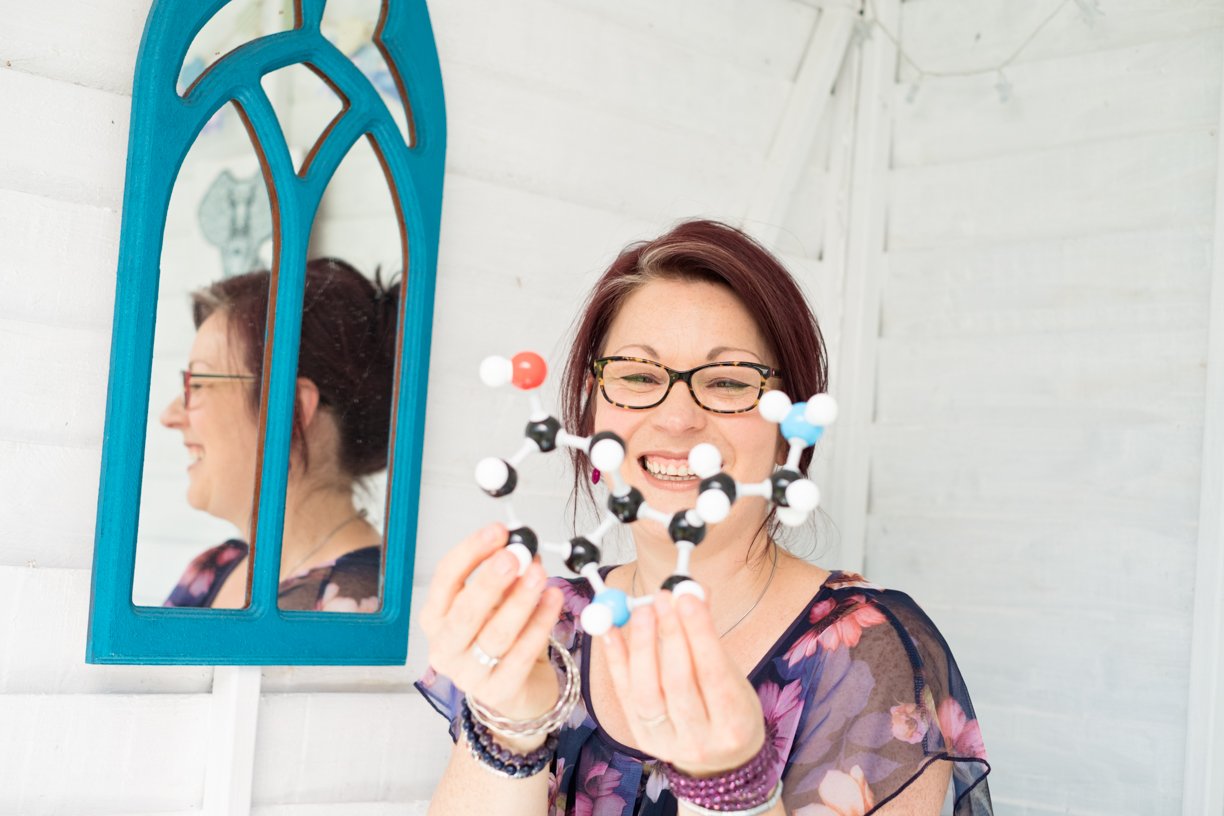 A smiling woman with glasses and dark red hair holding a molecular model in front of a white wall with a blue mirror.