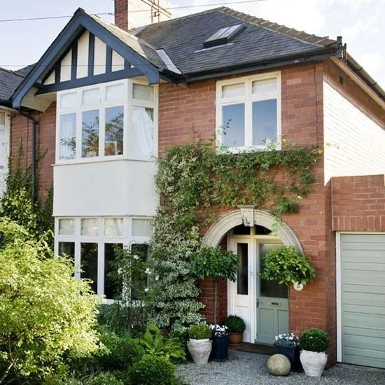 Front view of a brick house with a bay window and an arched door, surrounded by green plants and potted flowers.