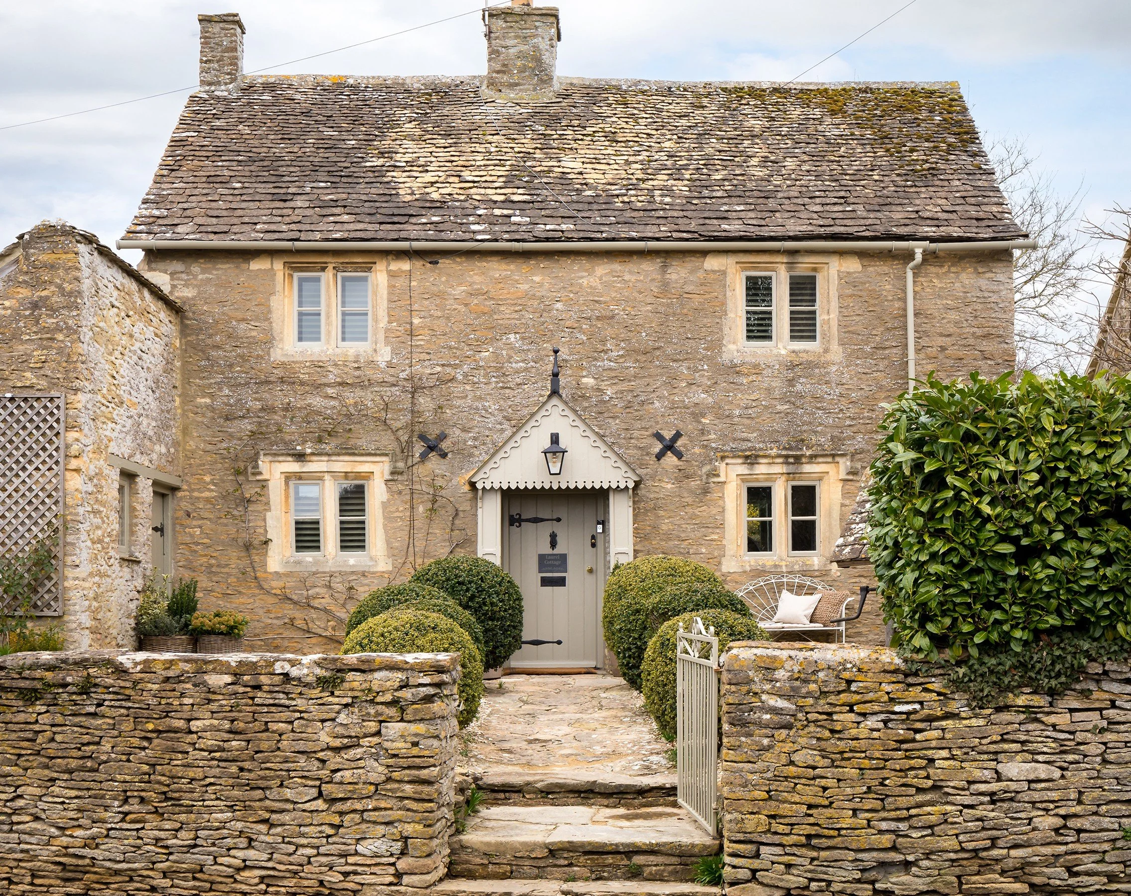 A stone cottage with a stone pathway leading to the front door, surrounded by a stone wall and greenery.