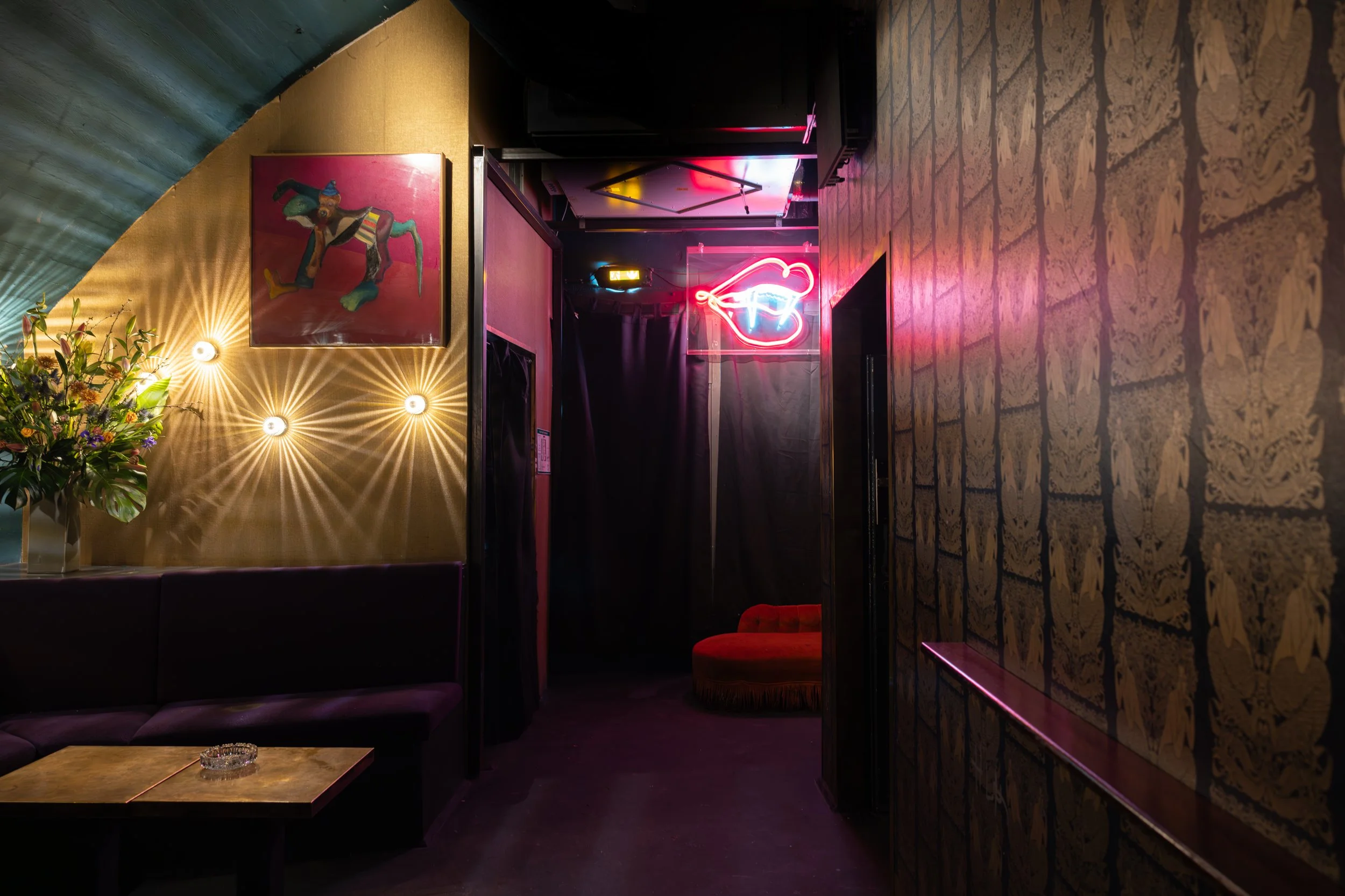 Innenaufnahme eines stilvollen Bars oder Lounges mit moderner Dekoration, Neonlicht in Form eines Hufeisens, Gemälde an der Wand, Blumen auf einem Tisch, Samtsofa und einer roten Sitzgelegenheit im Hintergrund.