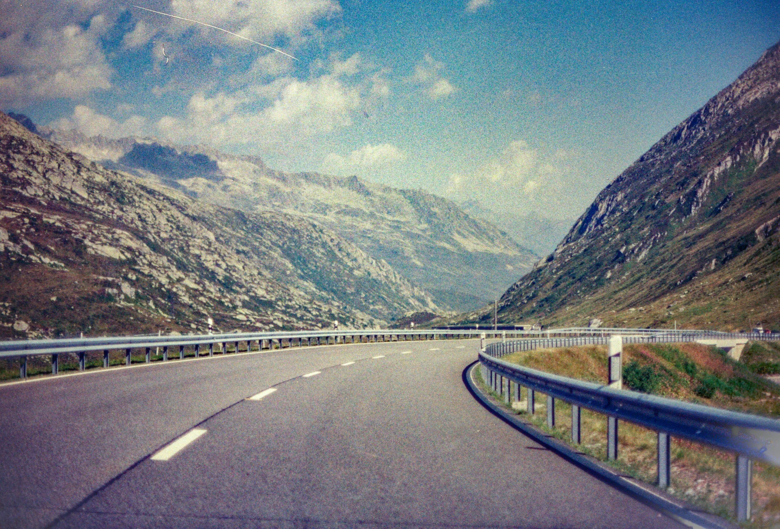 Kurvige Bergstraße in einer bergigen Landschaft mit hohen, steil abfallenden Felswänden, grünem Bewuchs am Straßenrand, ein blauer Himmel mit Wolken und Flugzeugspur am Himmel.