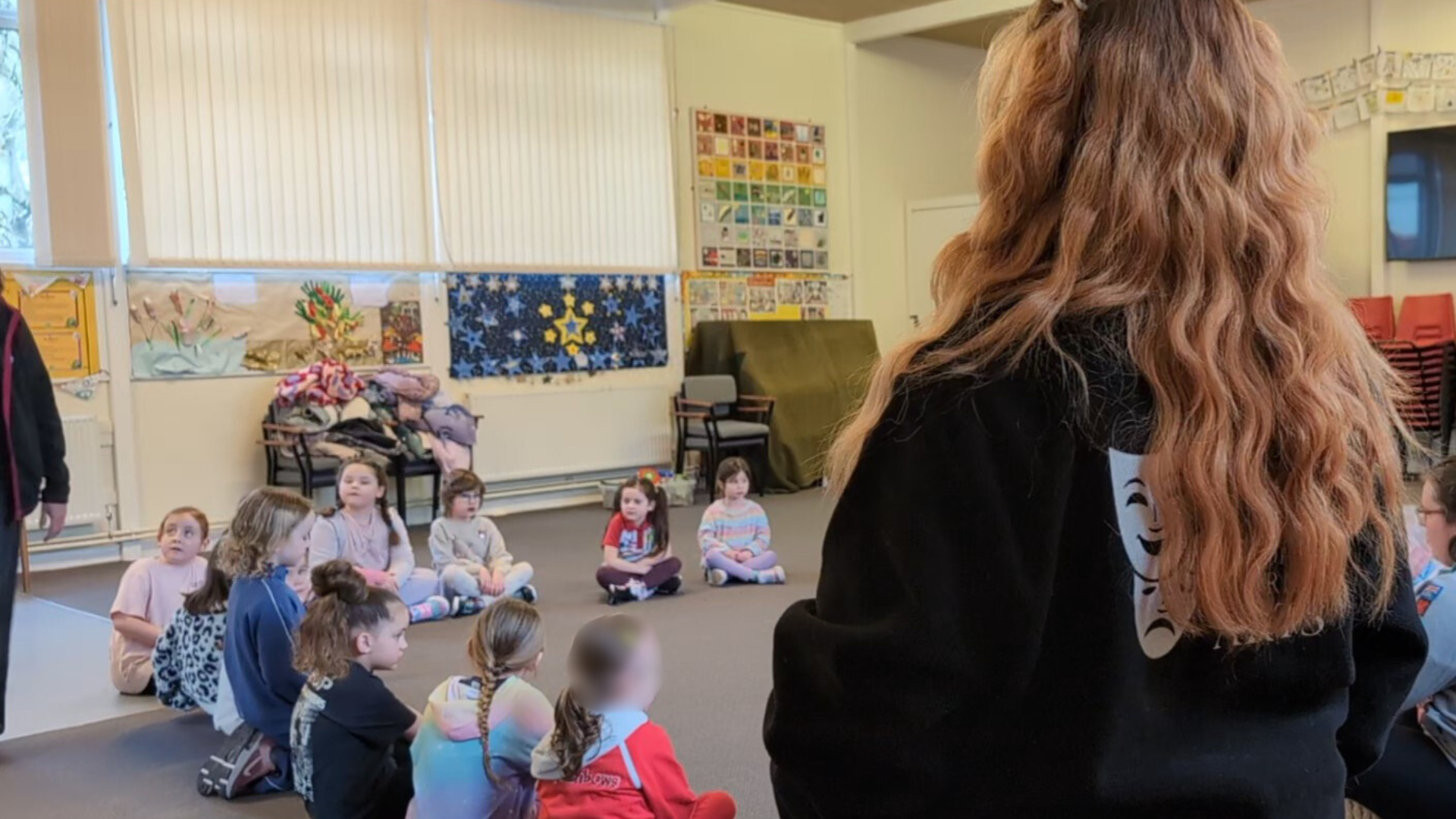 A classroom with children sitting on the floor and a woman standing in front. Decorations and artwork are on the walls, including stars and colorful drawings. The woman has long curly hair and is wearing a black hoodie with a white comedy and tragedy mask design on the back.