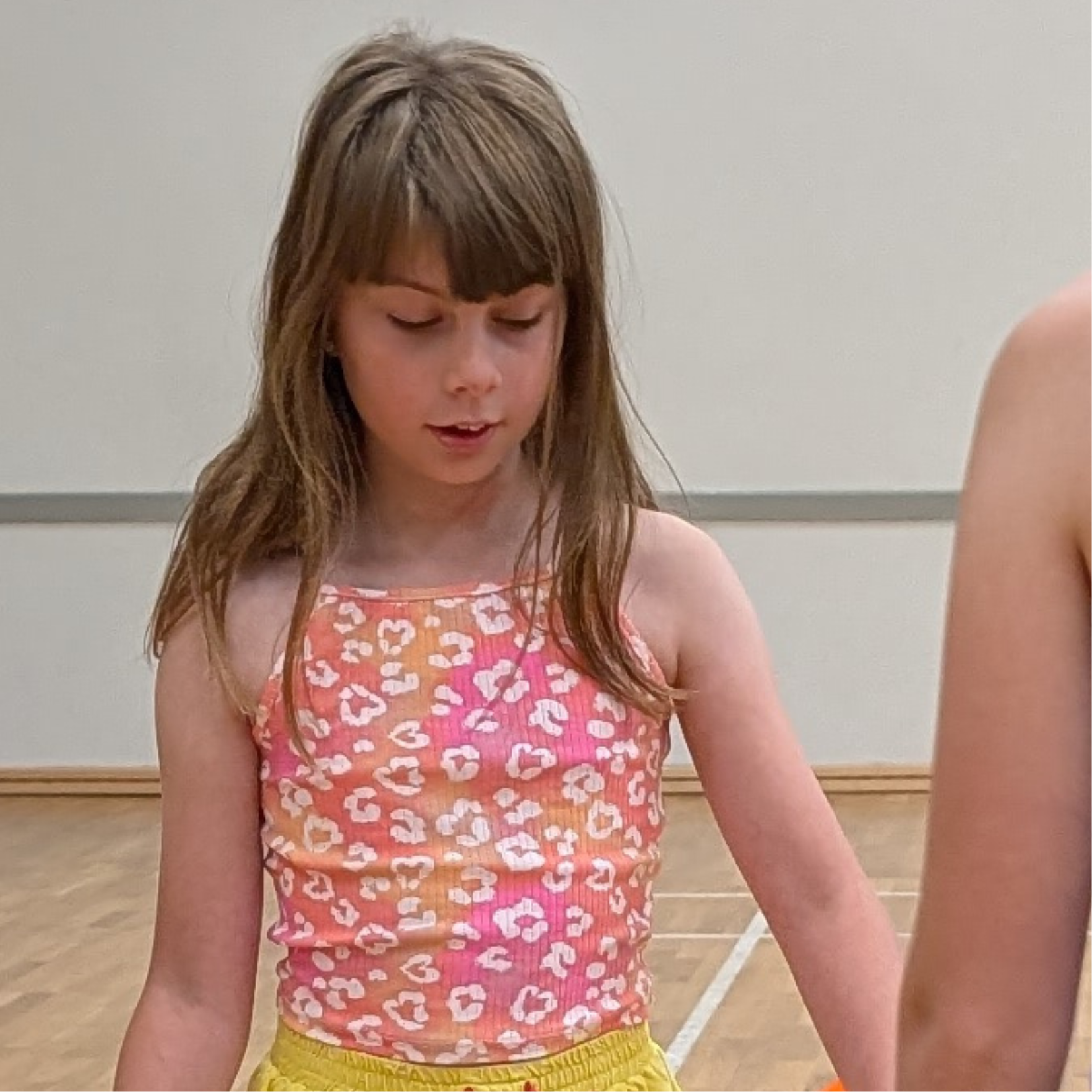 A young girl with long brown hair and bangs, wearing a pink and orange leopard print sleeveless top and yellow shorts, looking down while indoors.