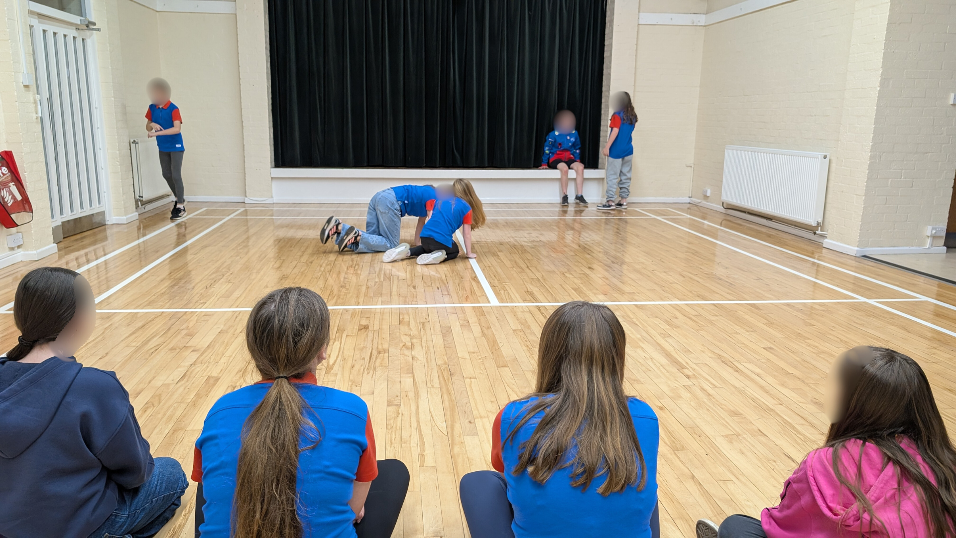 Children watching a play or performance with a stage and black curtain, some sitting on the floor and others performing.