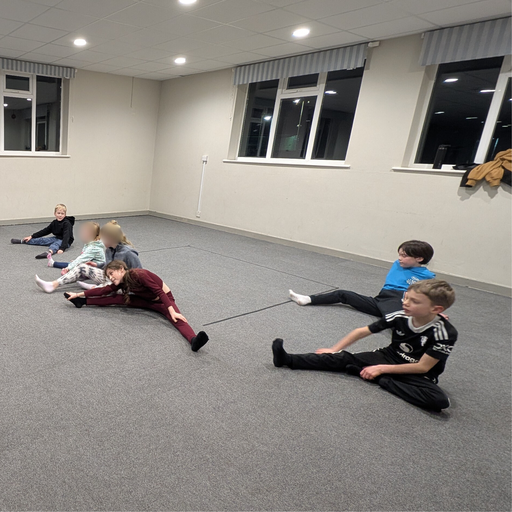 Children sitting on the floor in a spacious room, stretching with legs extended and arms reaching towards their feet, during a group activity.