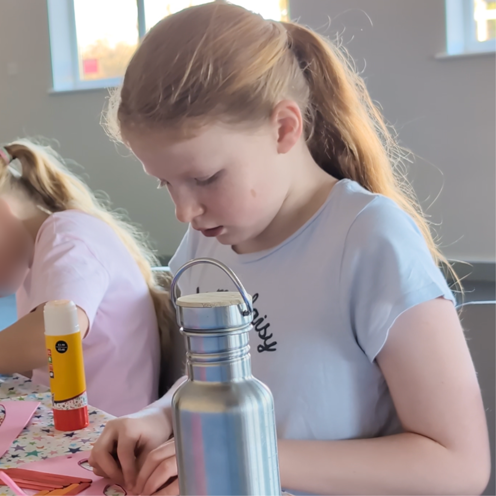 A young girl with red hair in a ponytail, wearing a white t-shirt, focused on a craft activity at a table with a pink tablecloth, nearby are glue sticks and paper. The scene is in a bright room with windows letting in natural light.