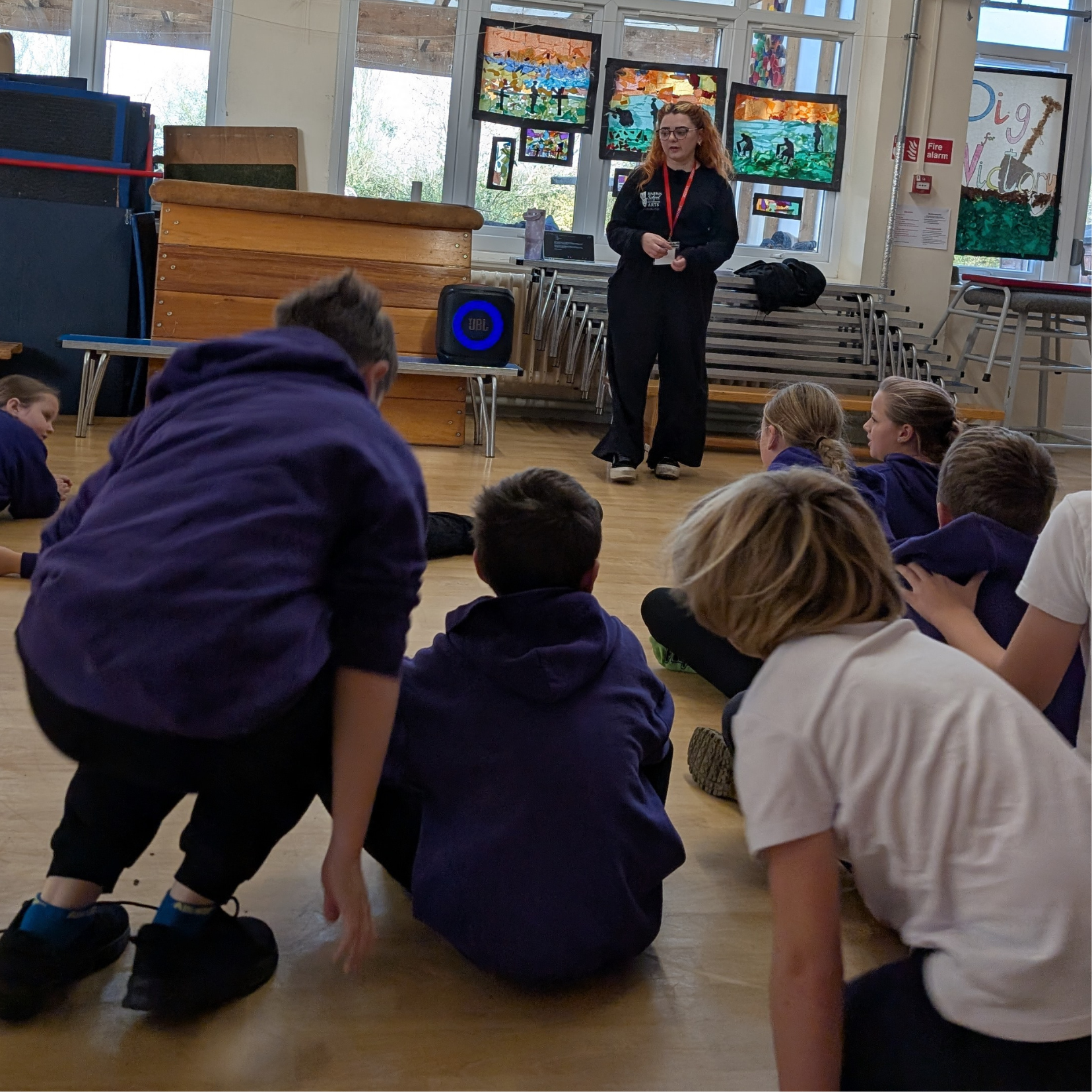 School classroom with children sitting on the floor listening to a female instructor standing in front of colorful artwork displayed on windows.