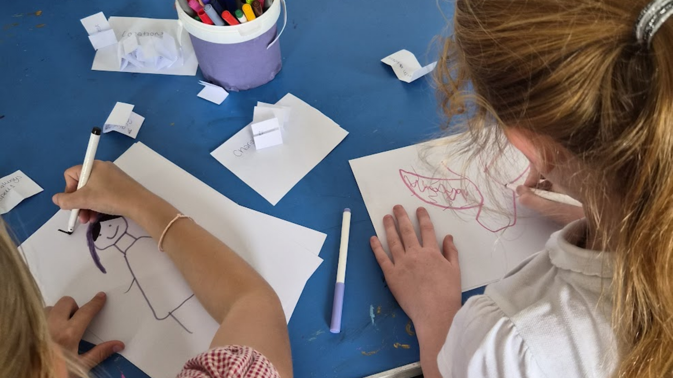 Two young girls sitting at a blue table, drawing characters with markers on white paper. One girl is drawing a girl, and the other is drawing a face with big teeth. There are scattered paper scraps and a container filled with colorful markers.