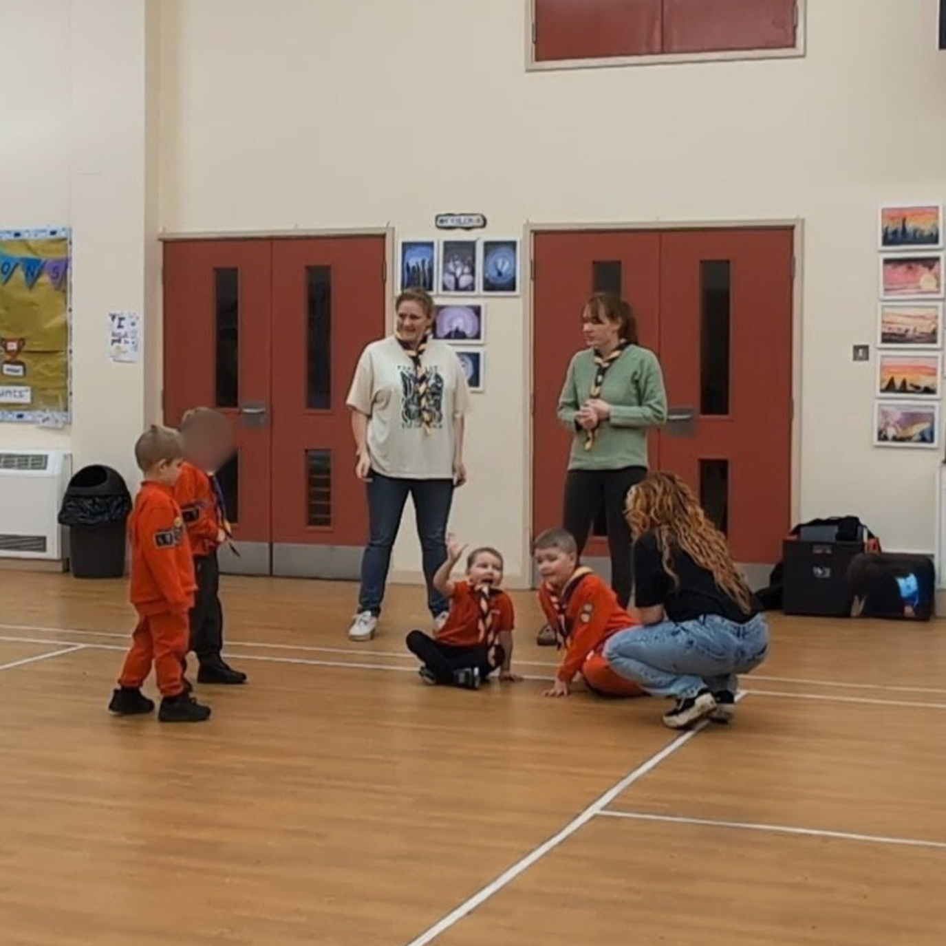 Children and adults in a gymnasium, with some children sitting and others standing, participating in a group creative activity.