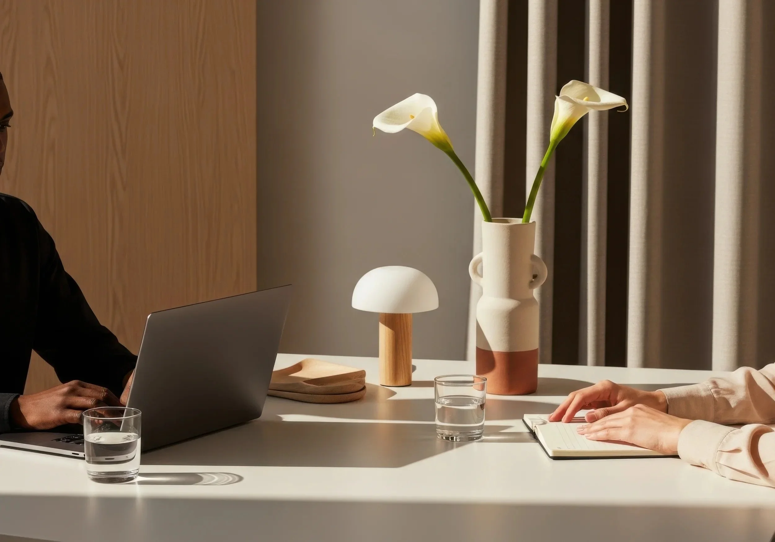 A minimalist office table with a laptop, two glasses of water, a small wooden table lamp, a vase with two white calla lilies, and a person taking notes on a notepad