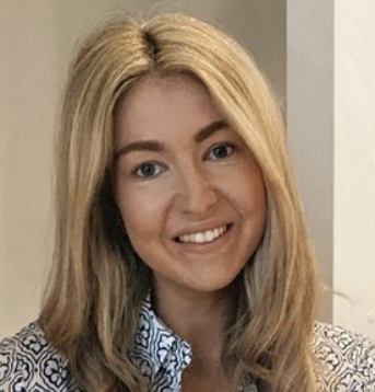A smiling woman with blonde hair and blue eyes, wearing a patterned blouse, standing indoors.